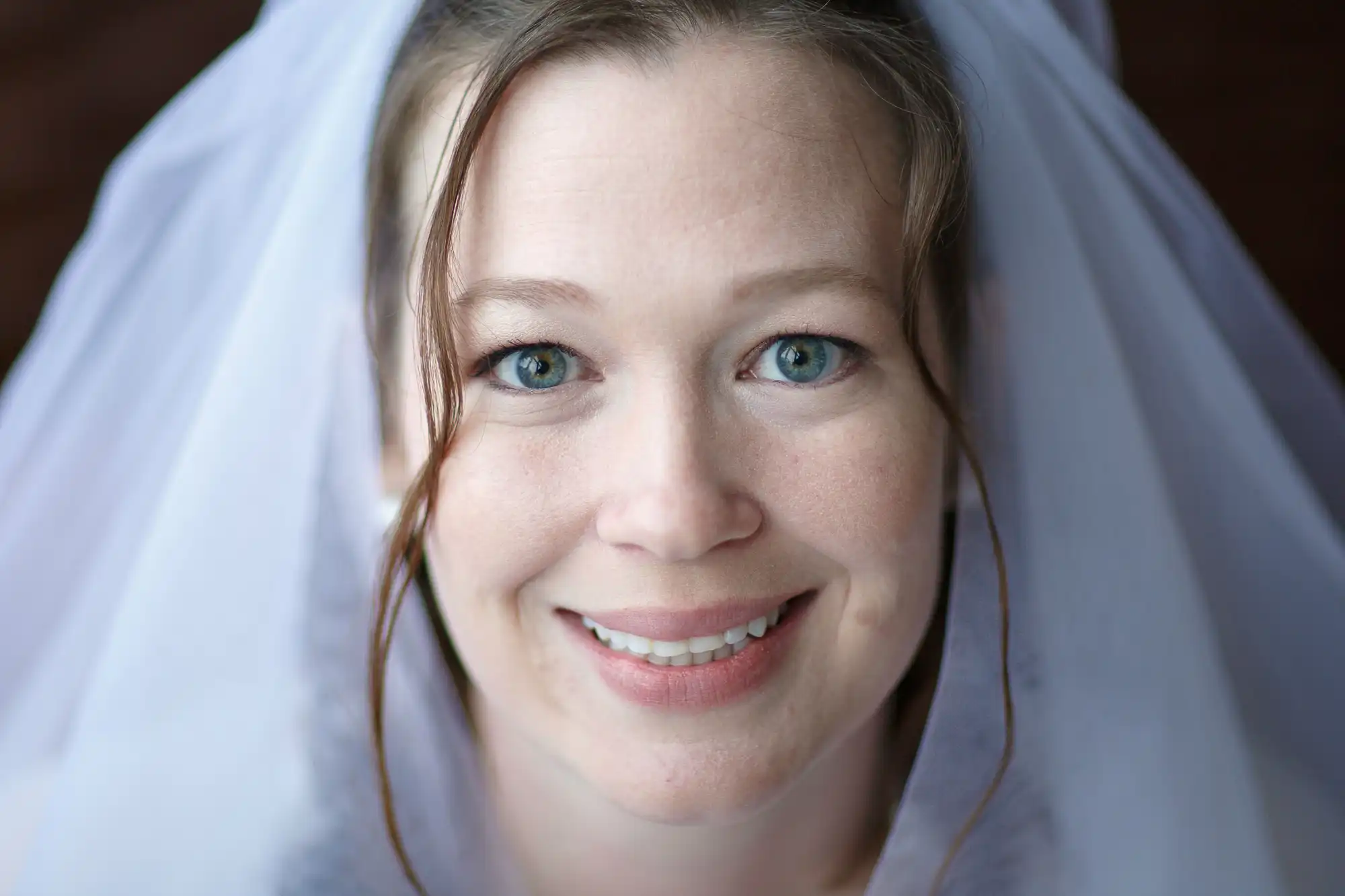 A close-up portrait of a smiling bride with bright blue eyes, draped in a white veil.