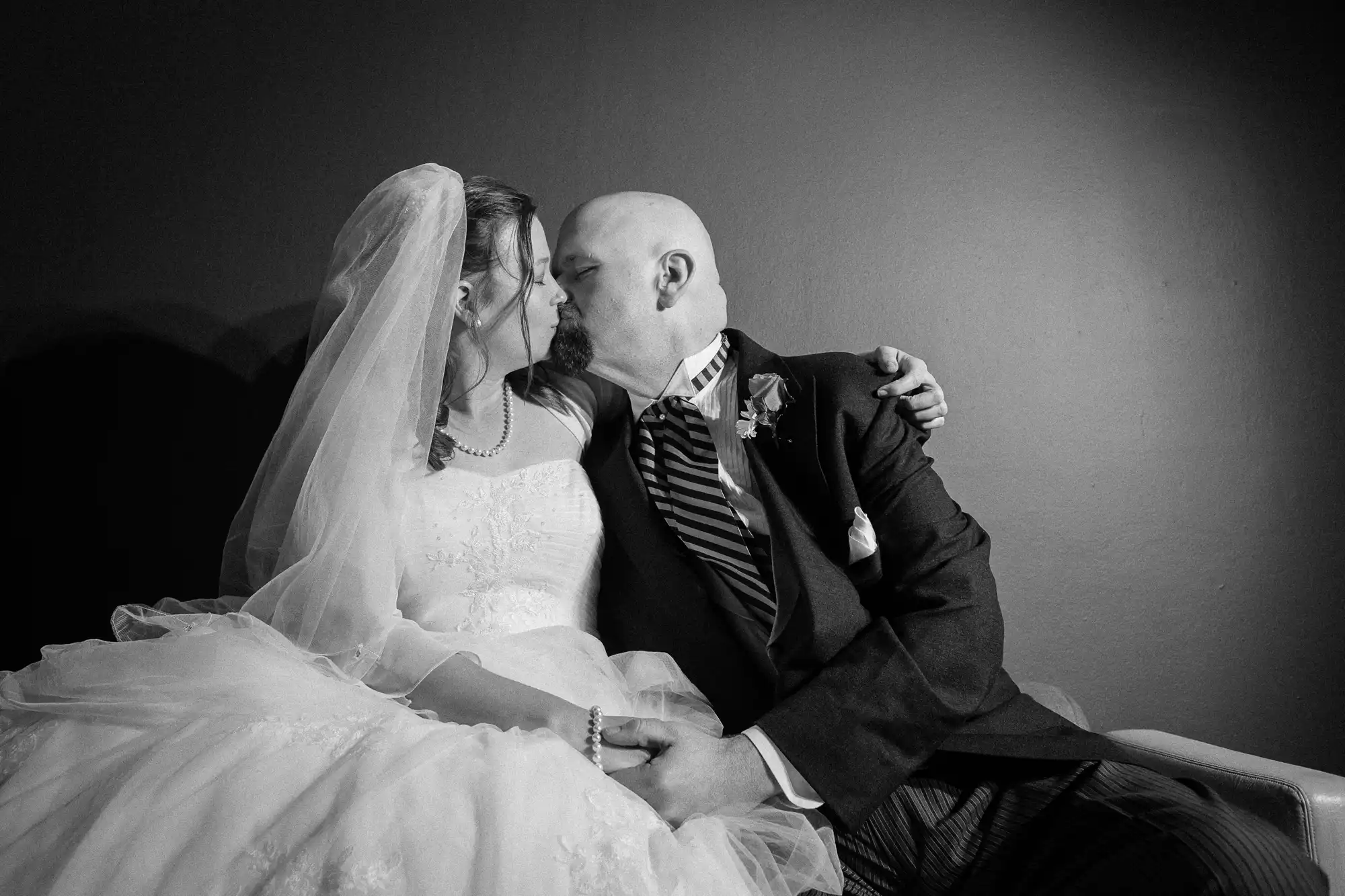 A bride and groom share a tender kiss, seated closely, in a black and white photograph.