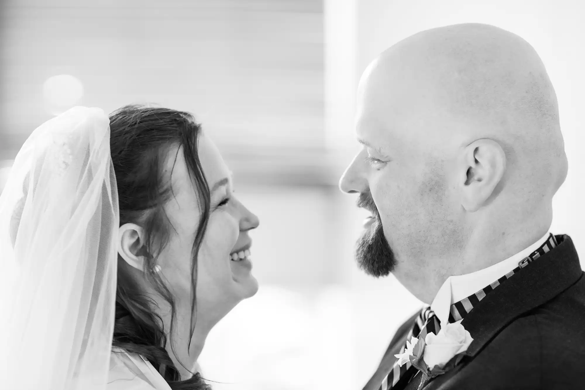 A bride and groom smiling at each other, both in wedding attire, captured in a close-up black and white photograph.