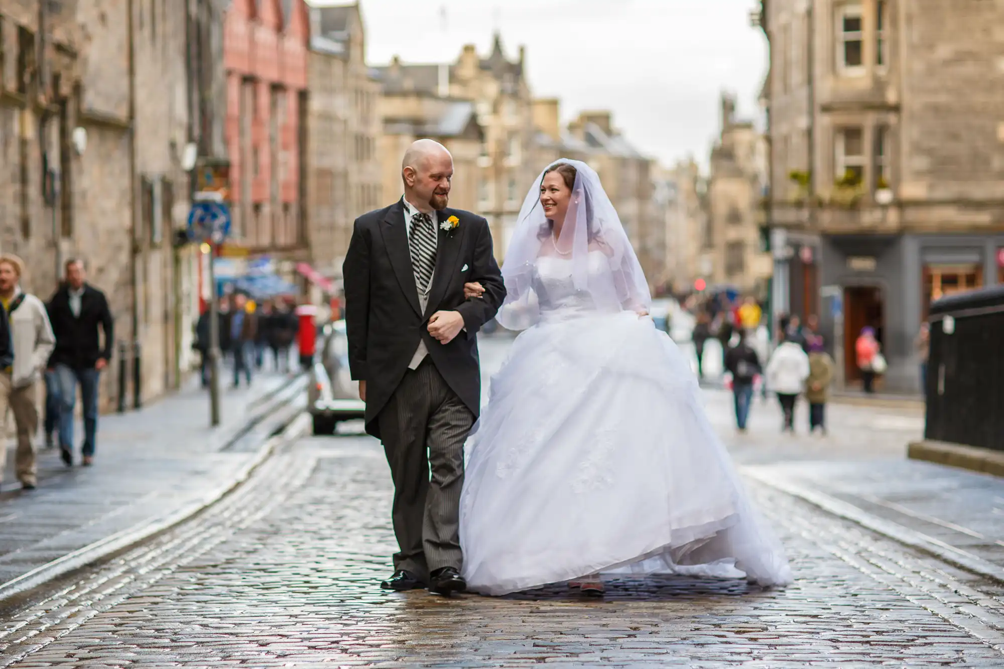 A bride and groom walk hand in hand on a cobbled street, smiling joyfully, with people and historic buildings in the background.
