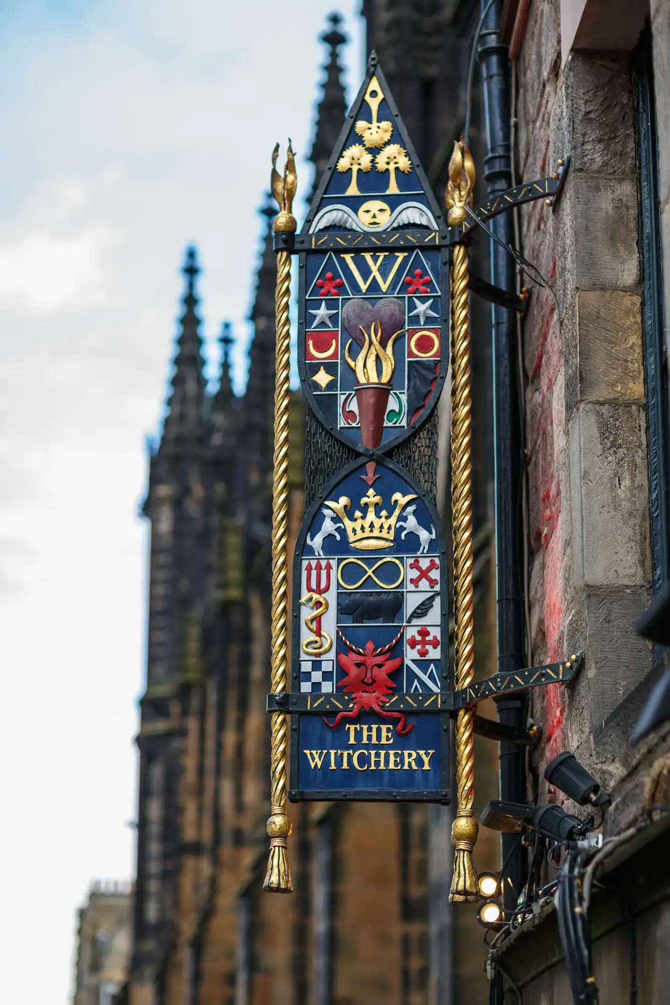 Ornate sign for "the witchery" featuring heraldic shields and symbols, with a gothic tower in the soft-focus background.