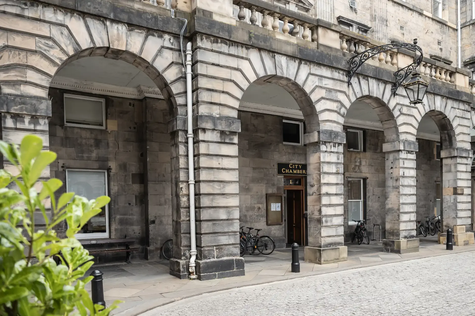 wedding photographer at Edinburgh City Chambers A stone building labeled "City Chambers" features arched entrances, a lantern light fixture, and a few parked bicycles. A cobblestone street and some greenery are visible in the foreground.