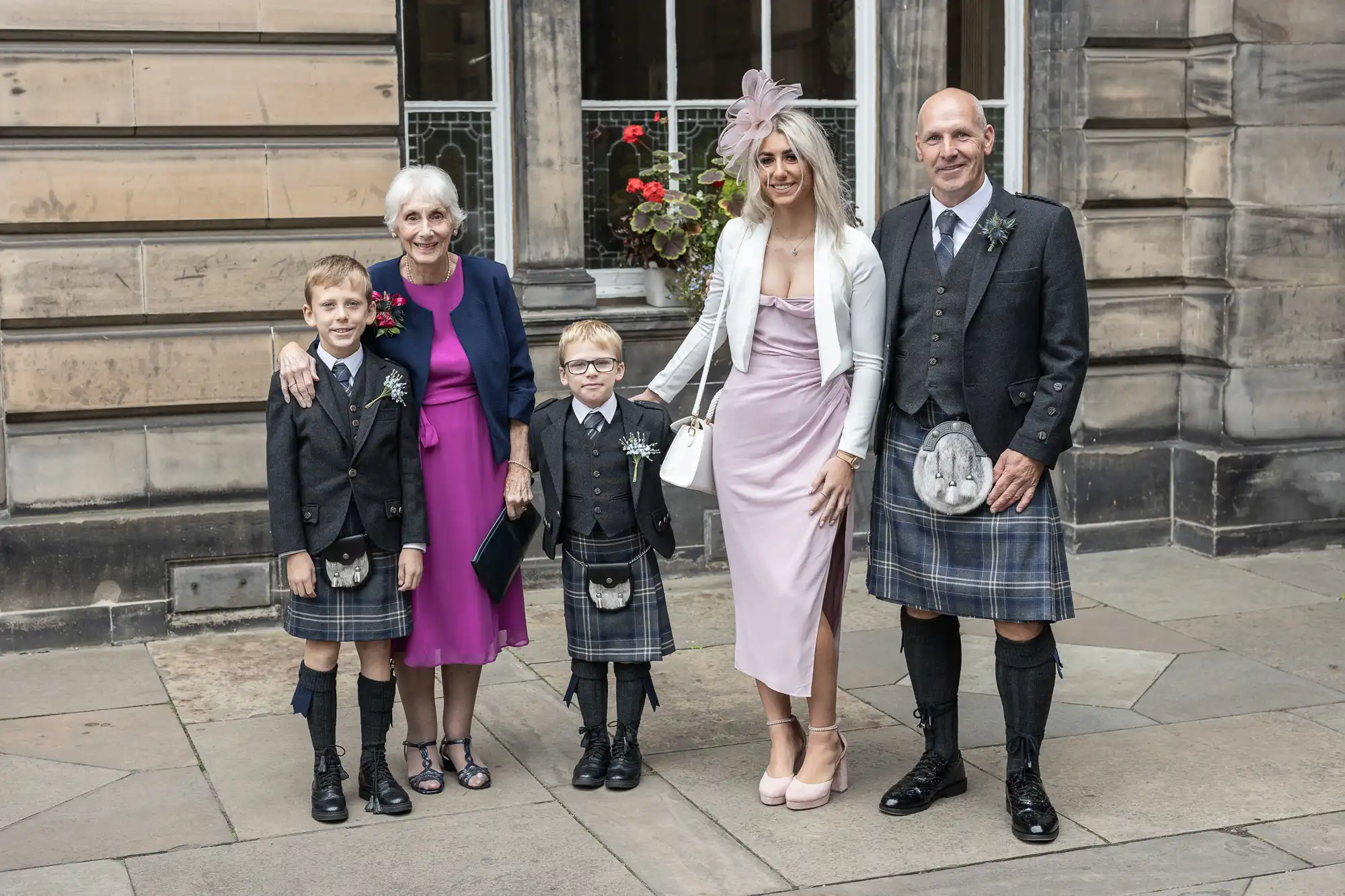 A family dressed in formal attire, with two men and two young boys in kilts, an elderly woman in a purple dress, and a woman in a pink dress and white jacket, standing outside a stone building.