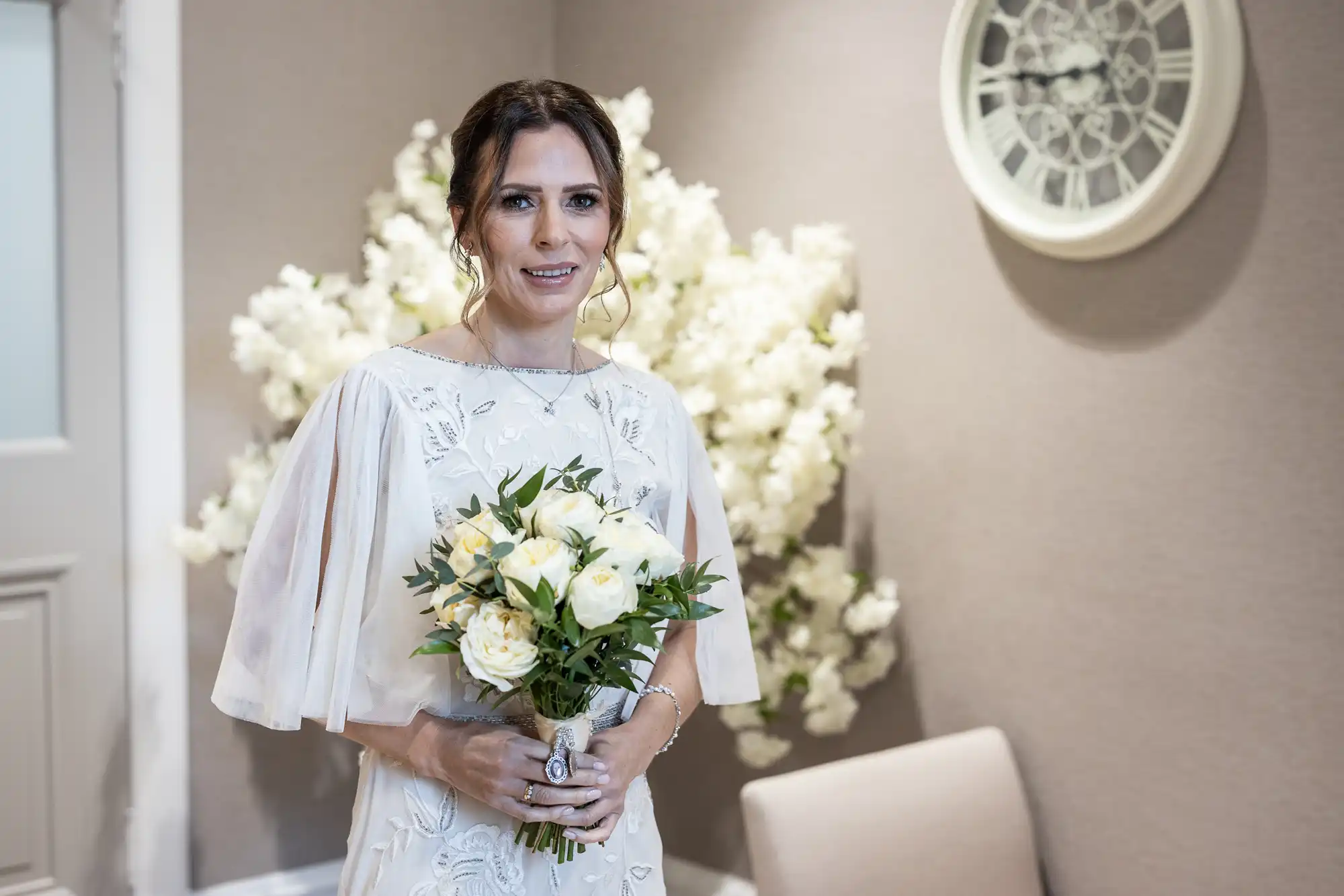 A woman wearing a white dress holds a bouquet of white flowers, standing indoors with a wall clock and white floral decorations in the background.