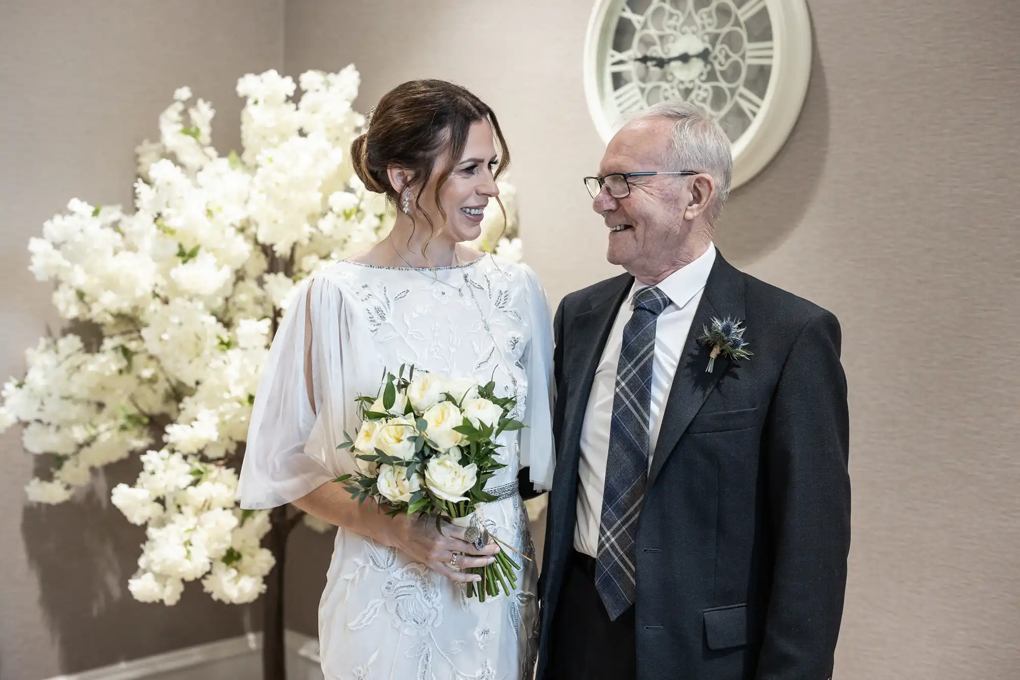 A smiling woman in a white dress holding a bouquet of white roses stands next to an older man in a suit and tie. They are standing in front of a flowering tree and a wall clock.