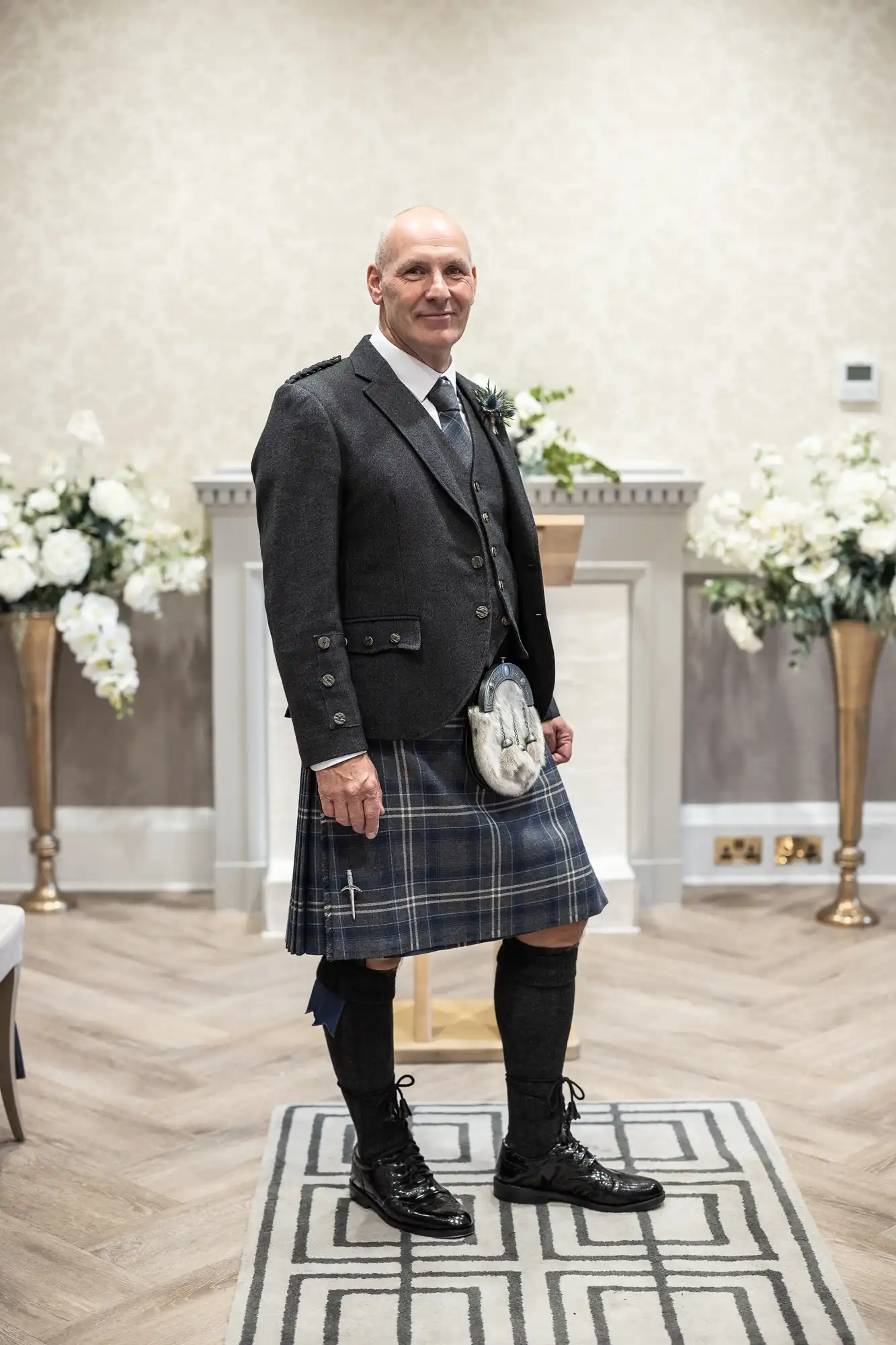 A man in formal Scottish attire, including a kilt and sporran, stands in a room with floral decorations and wood flooring.