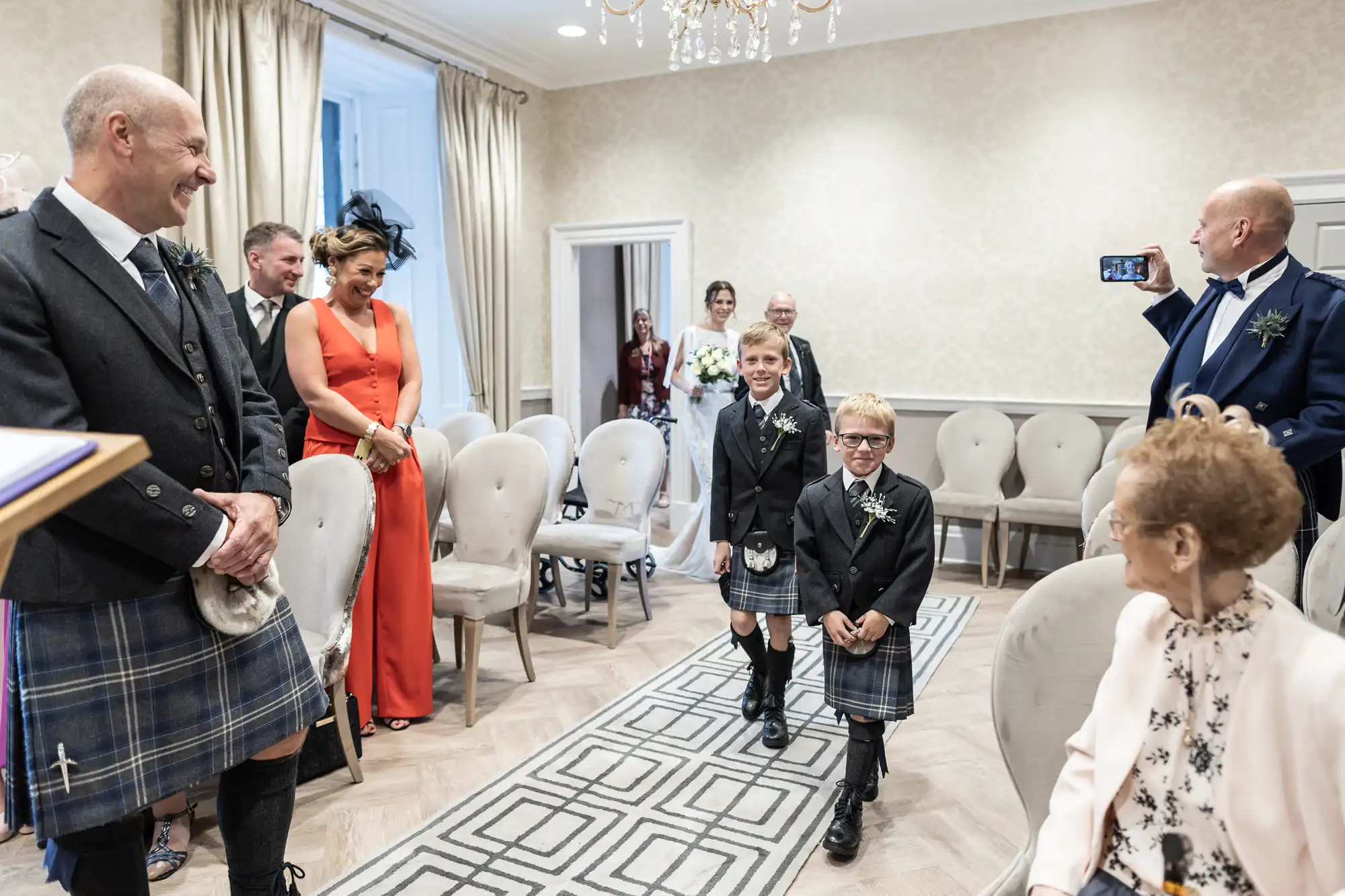 Two young boys in kilts walk down an aisle at a wedding ceremony. Guests, some in formal attire, smile and watch. One man captures the moment on his phone.