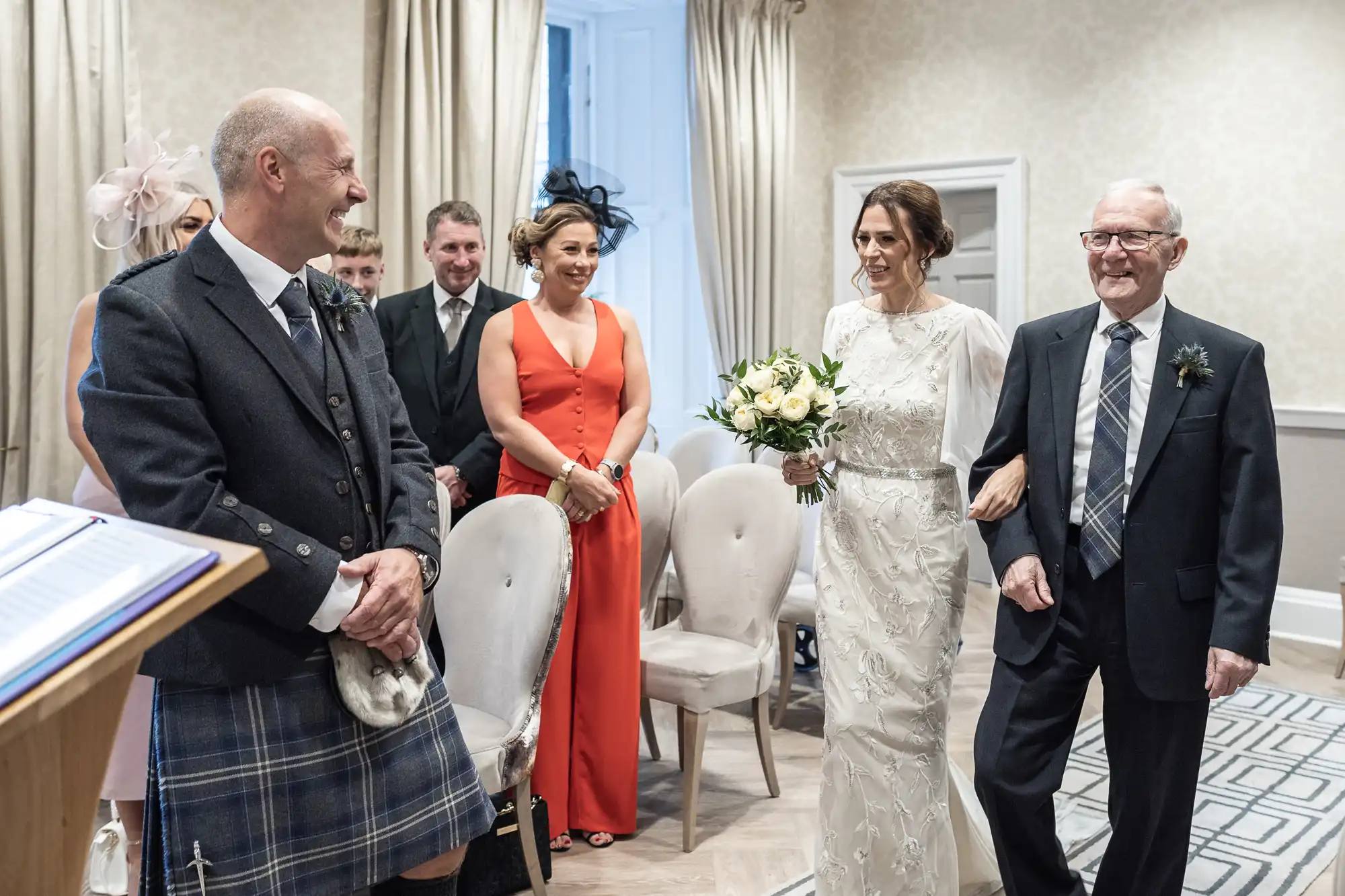 A bride, holding a bouquet, walks down the aisle with an older man. The groom, in a kilt, looks on happily. Wedding party members stand and watch.