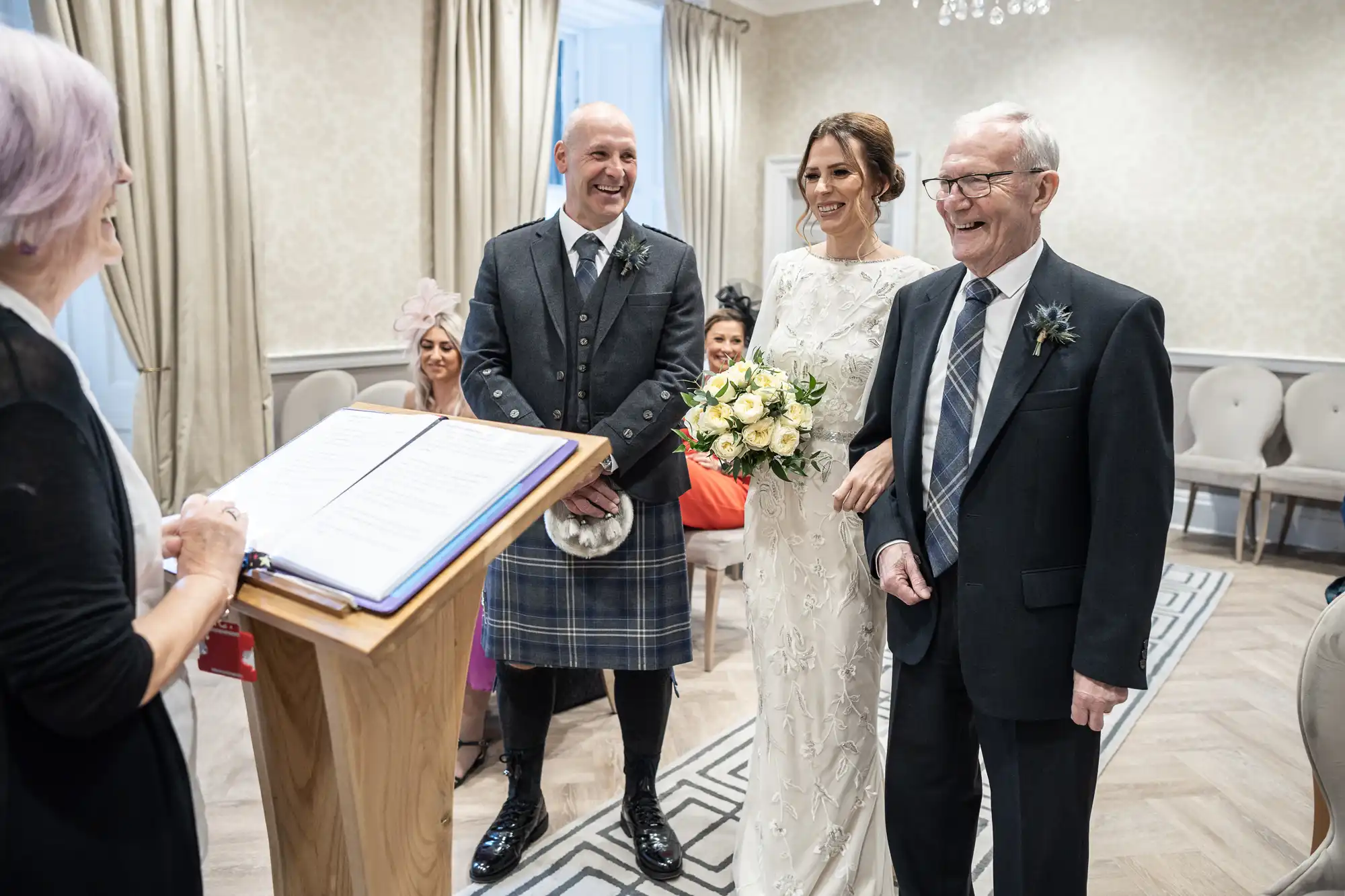 A bride and groom stand next to an elderly man during a wedding ceremony. The groom wears a kilt and the bride holds a bouquet. An officiant is reading from a book at a podium. Guests are seated behind.