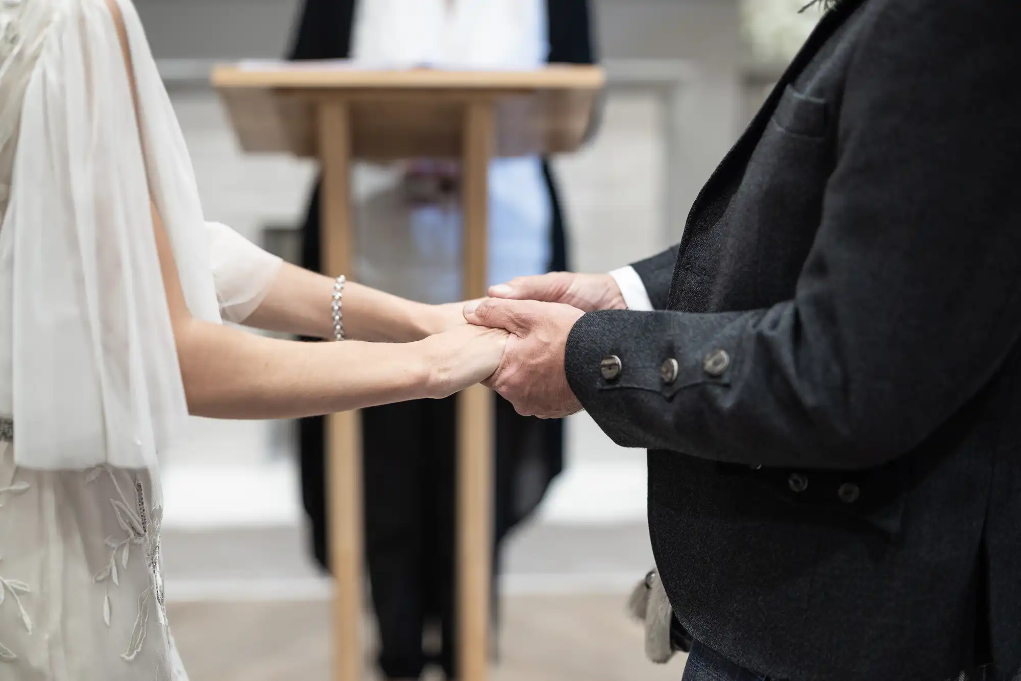 A bride and groom hold hands during a wedding ceremony, with an officiant standing at a wooden lectern in the background.
