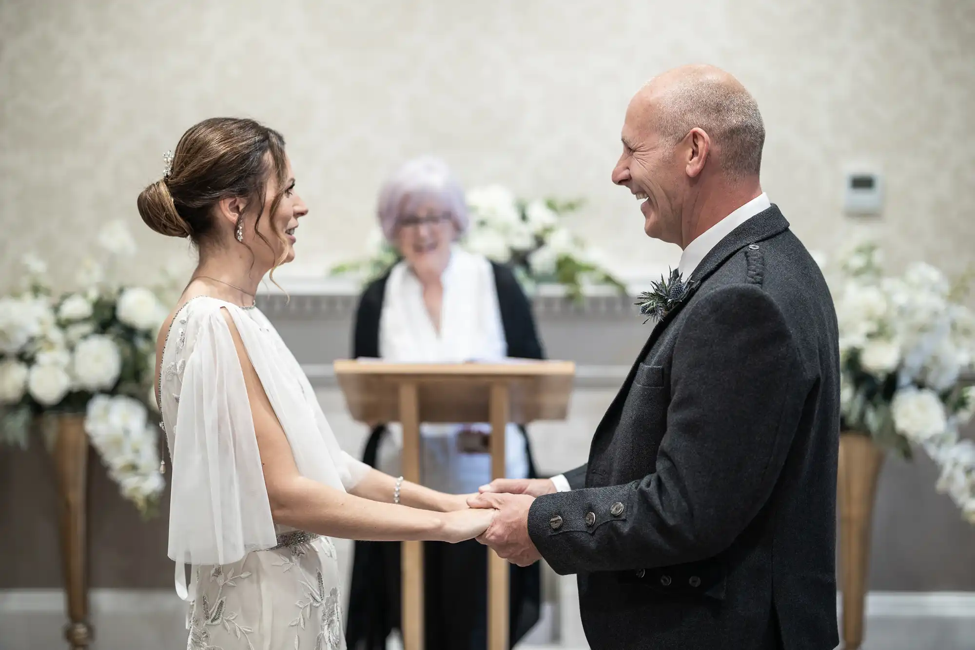 A couple holds hands and smiles at each other during their wedding ceremony, with an officiant standing behind them. The room is decorated with white flowers.