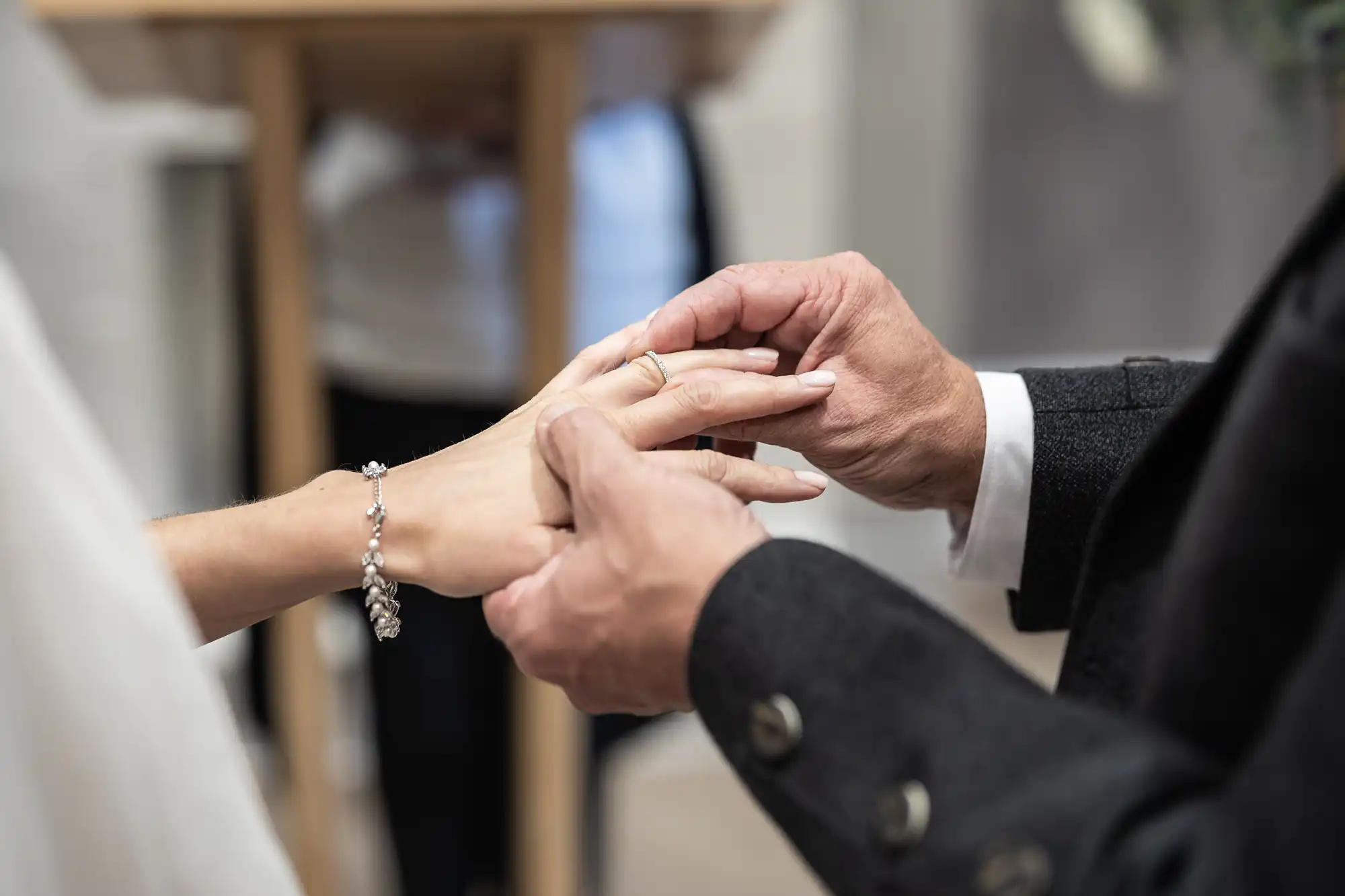 Close-up of a person placing a ring on another person's finger during a ceremony. The focus is on their hands, with one person wearing a bracelet and the other in a dark suit.
