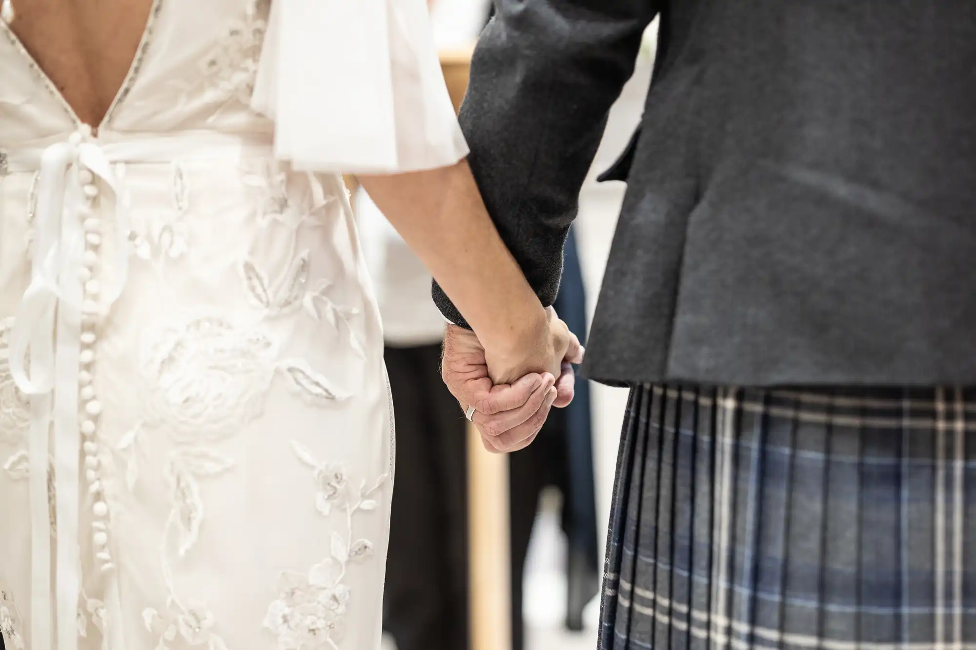 A couple holds hands, wearing a white wedding dress with floral embroidery and a dark suit with a plaid kilt, standing side by side.