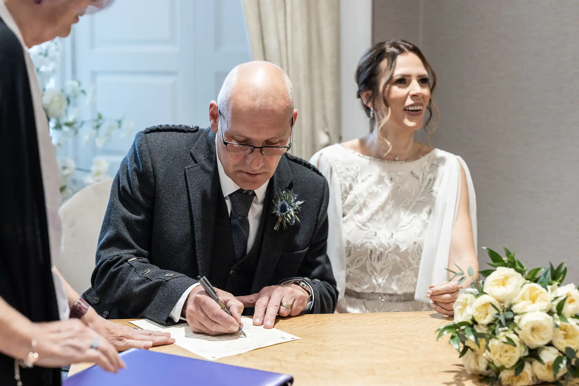 A man in a suit signs a document at a table beside a smiling woman in a white dress holding a bouquet of yellow roses. Another person holds a blue folder in the foreground.