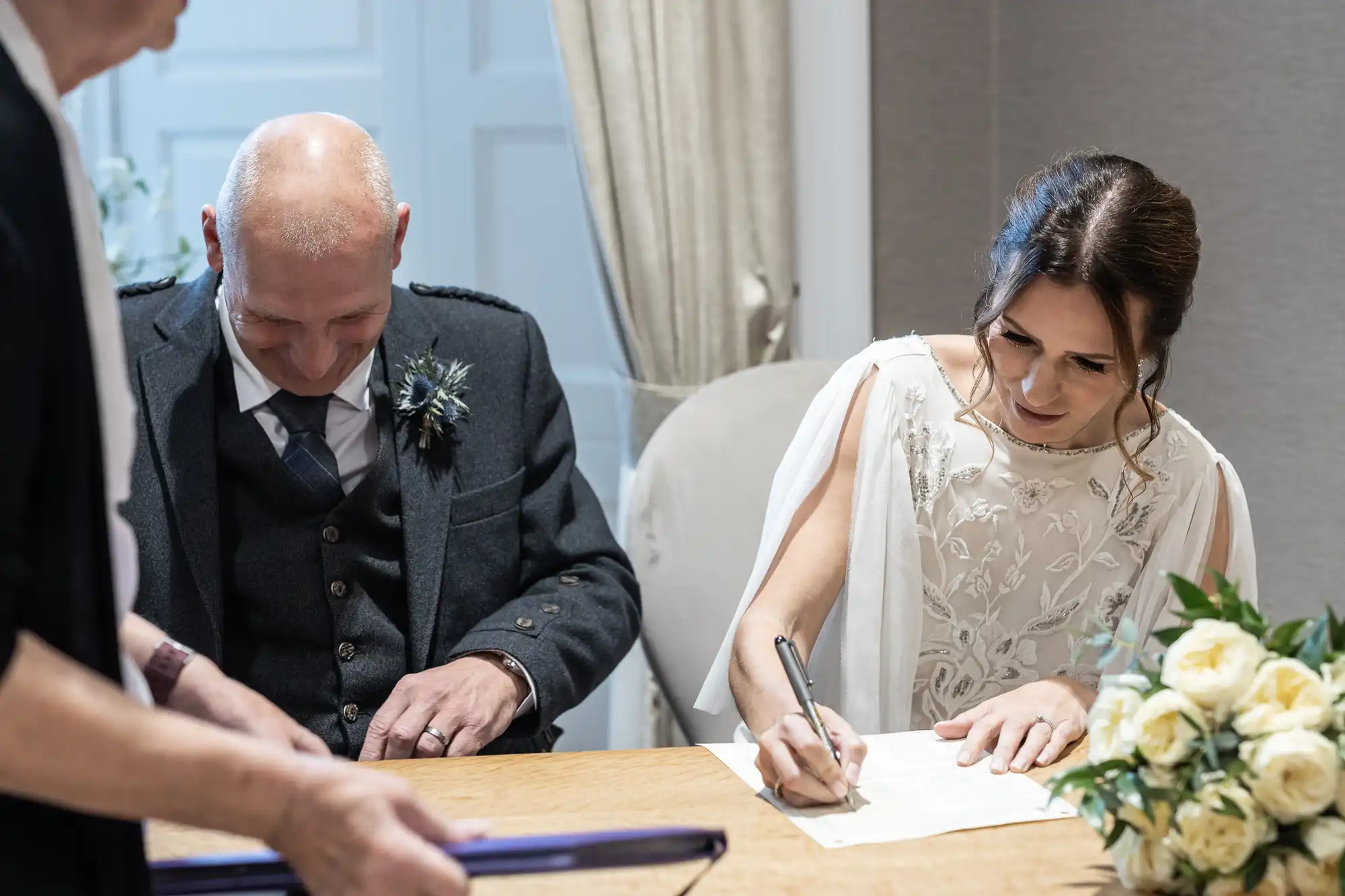 A bride signs a document at a wooden table while a bald man in formal attire sits beside her, smiling. Another person stands in the foreground. A bouquet of white roses sits on the table.