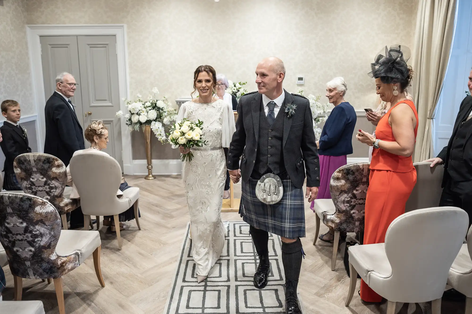 A bride and a man in a kilt walk down the aisle in a small, well-lit room with guests seated on both sides. The bride holds a bouquet and wears a white dress. The man wears traditional Scottish attire.