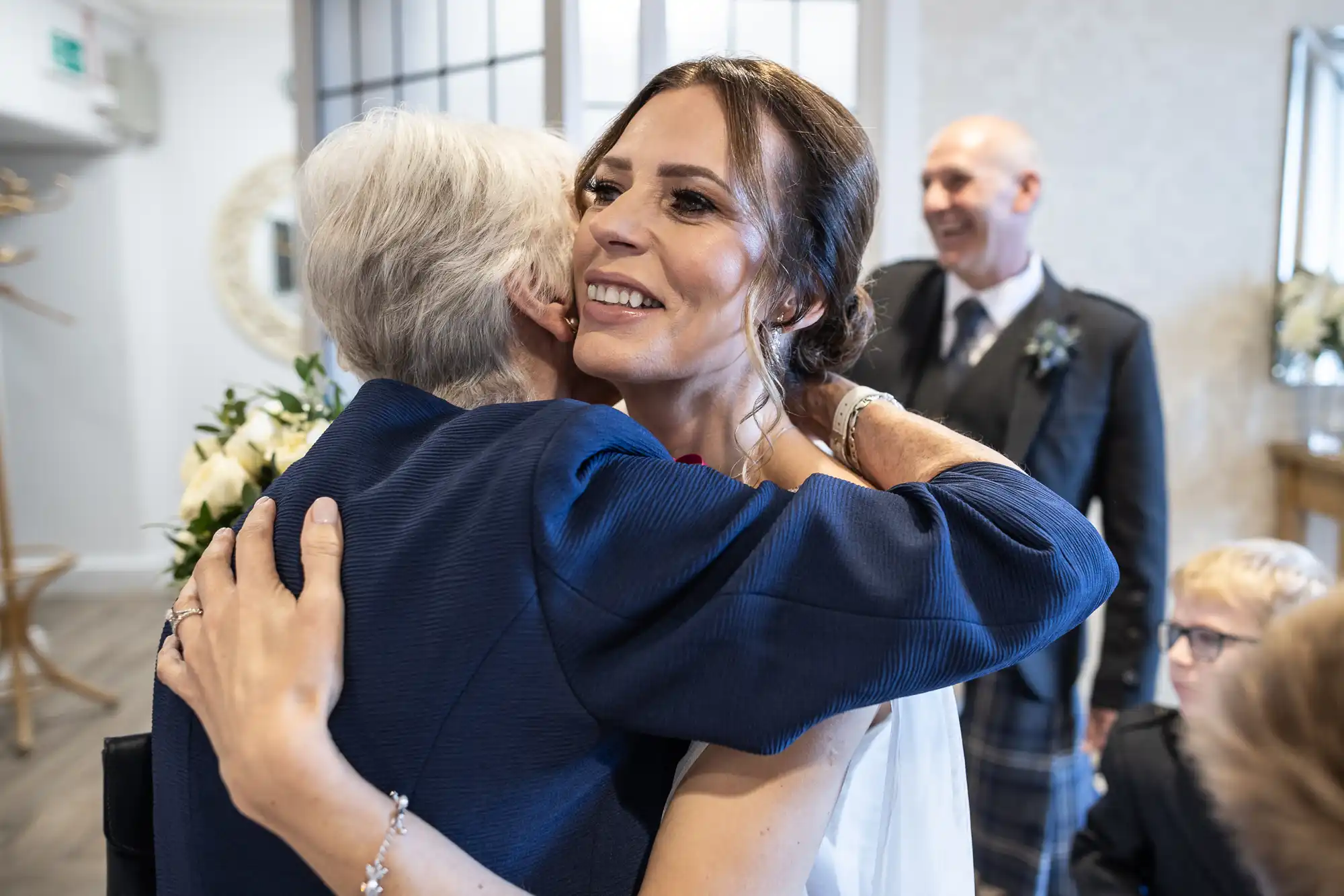 A bride in a white dress is hugging an older lady in a blue outfit. A man in a suit stands smiling in the background.