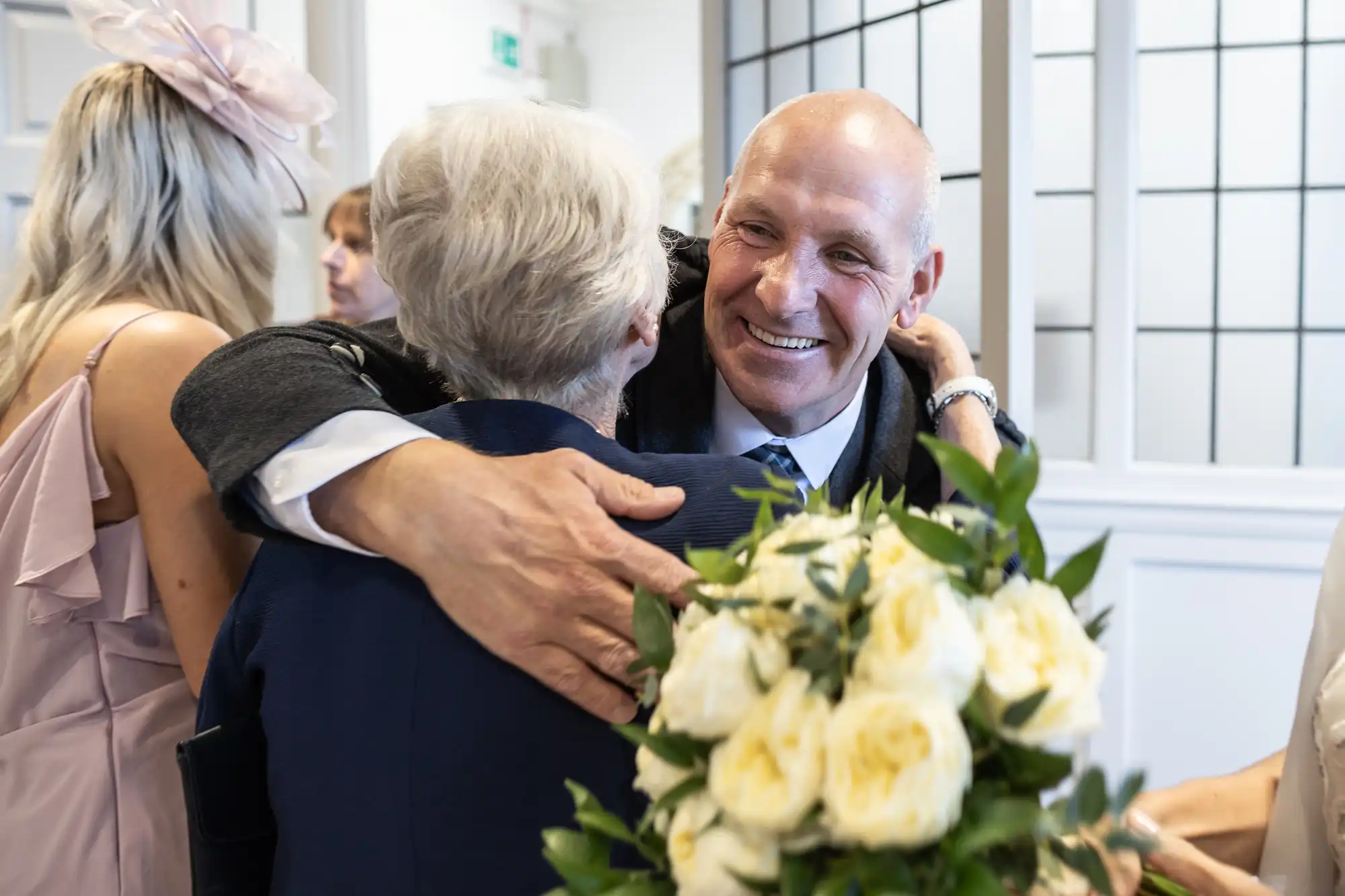 A man in a suit smiles warmly as he hugs an older woman. A bouquet of white flowers is in the foreground. Other people are in the background.
