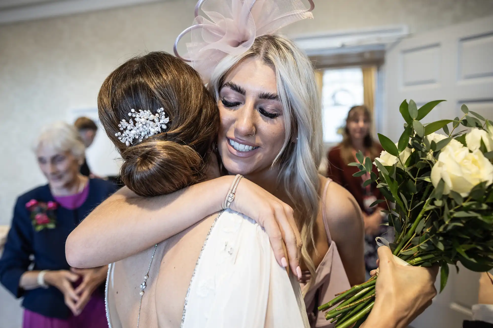 A woman with a floral hairpiece hugs another woman holding a bouquet of white roses in a well-lit room. An elderly woman in the background watches them.