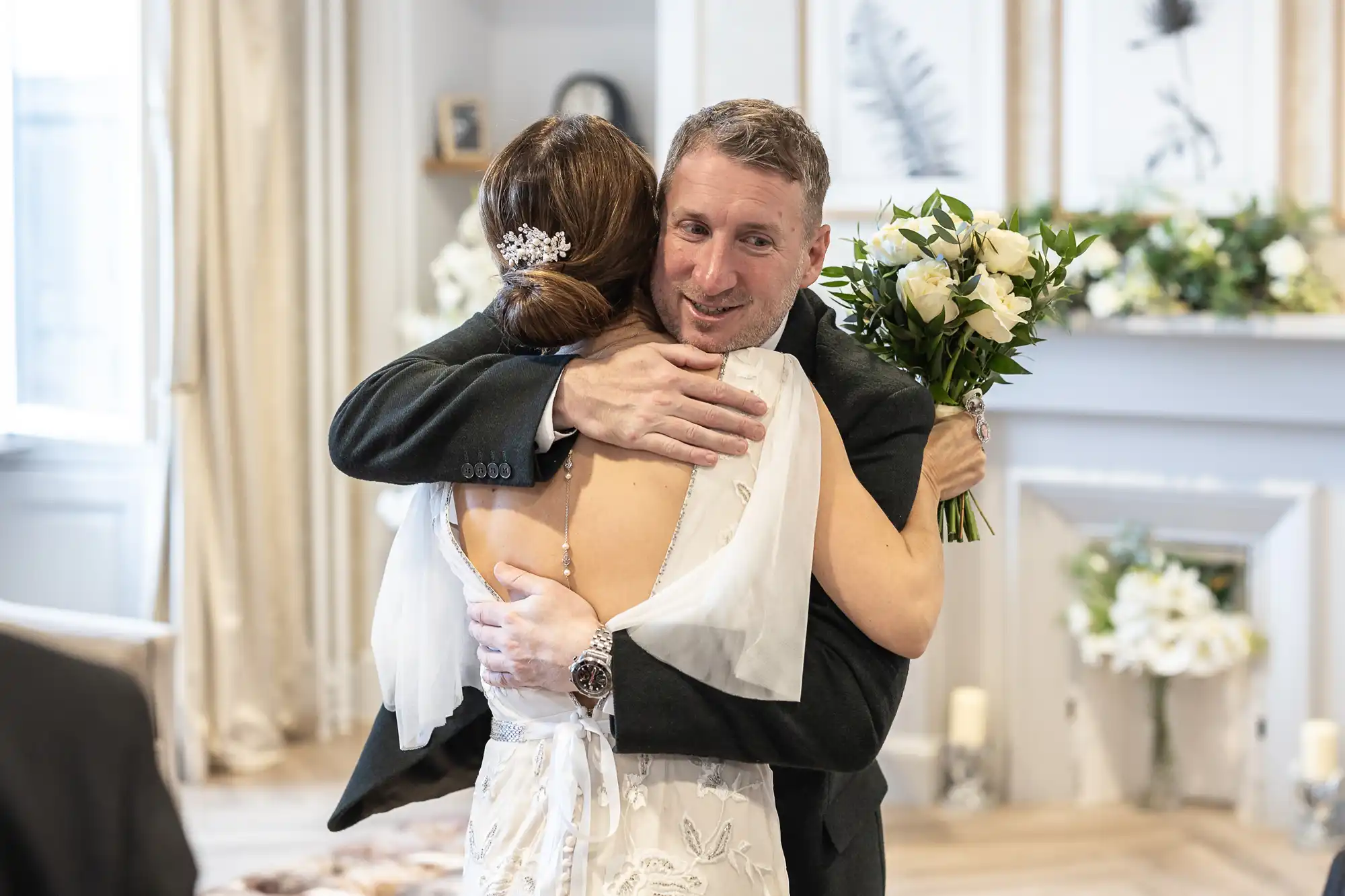 A man in a suit hugs a woman in a white dress holding a bouquet of flowers in a warmly decorated room.