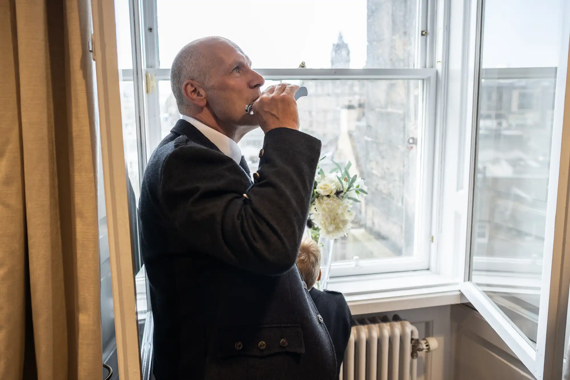 A man in a suit drinking from a flask while looking out a window with a cityscape view. A vase with flowers and a radiator are visible in the room.