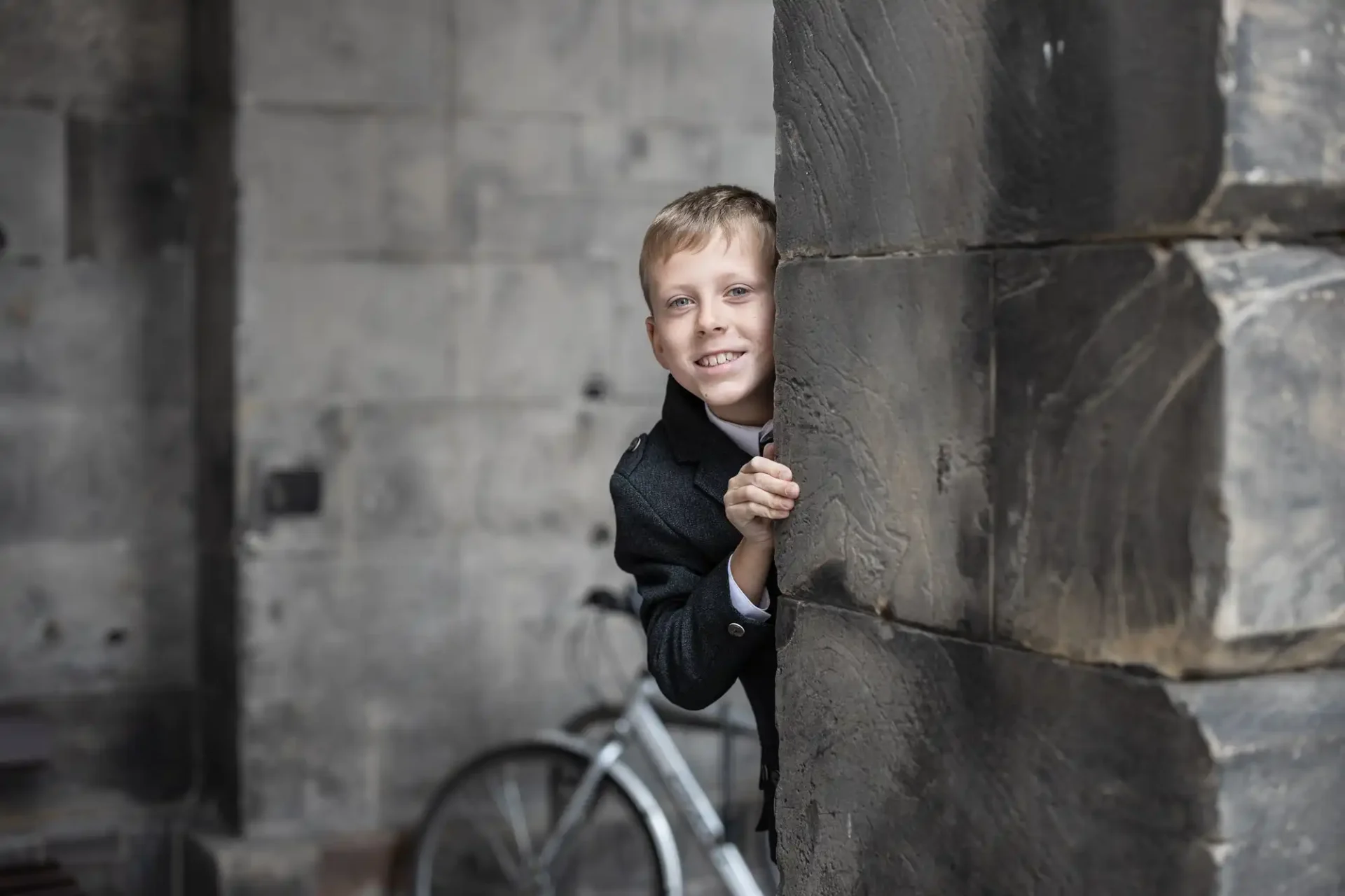 documentary style wedding photography example: A boy in a gray coat smiles as he peeks around a stone column. A bicycle is partially visible in the background.