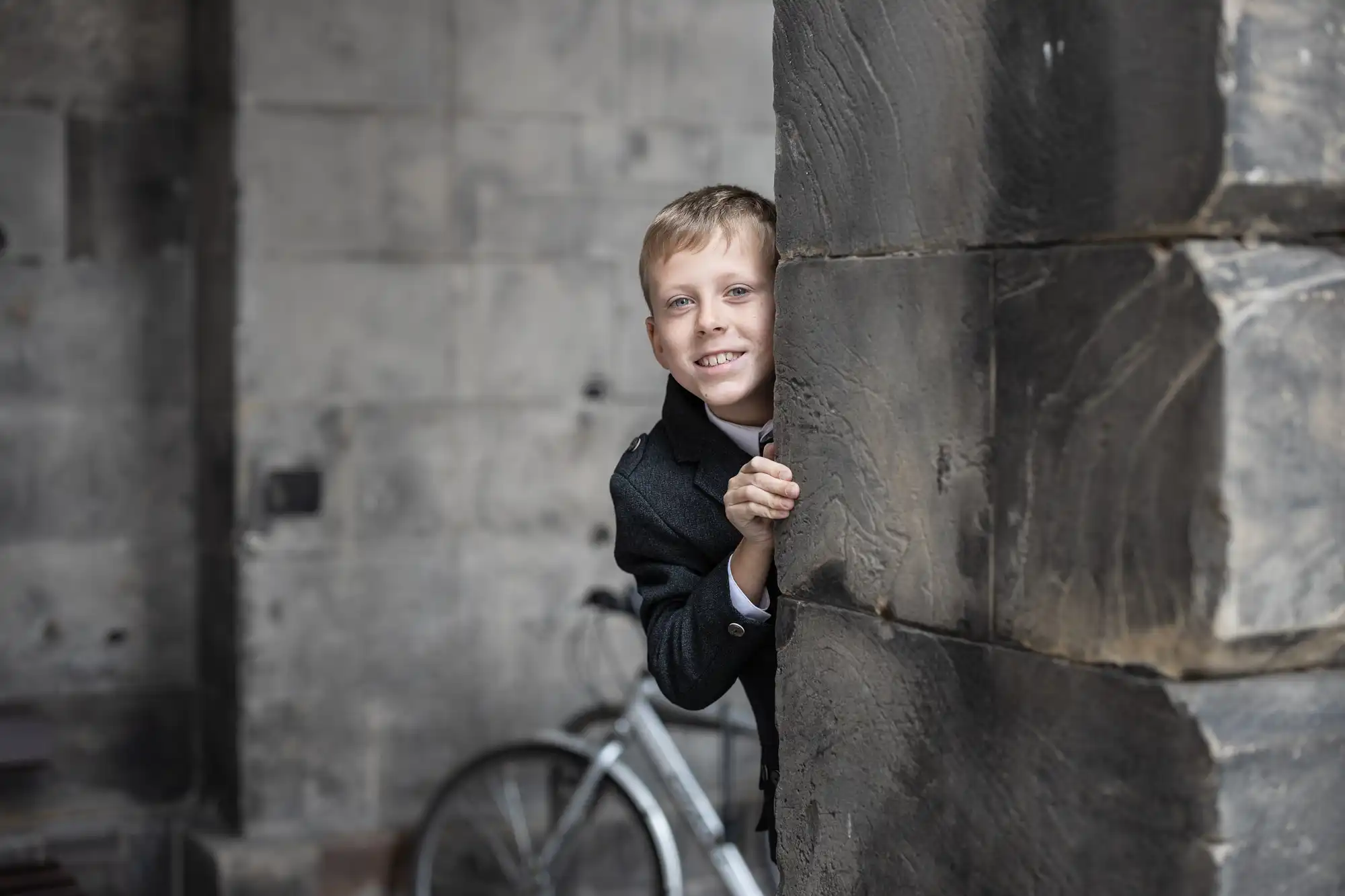 A boy in a gray coat smiles as he peeks around a stone column. A bicycle is partially visible in the background.