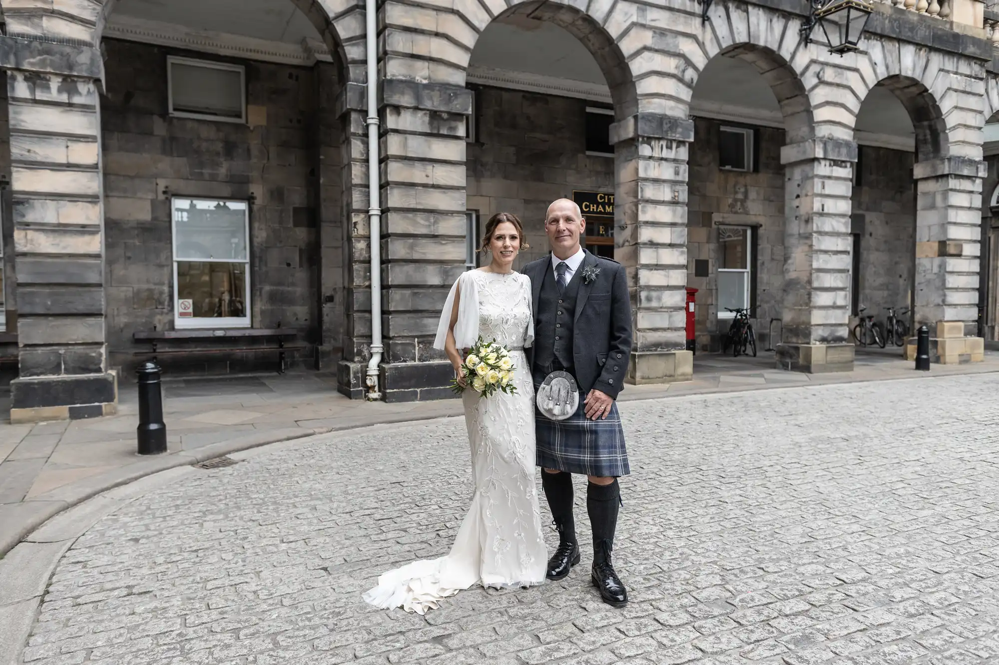 A bride in a white dress and a groom in a kilt stand side by side on a cobblestone street in front of a historic stone building with arches.