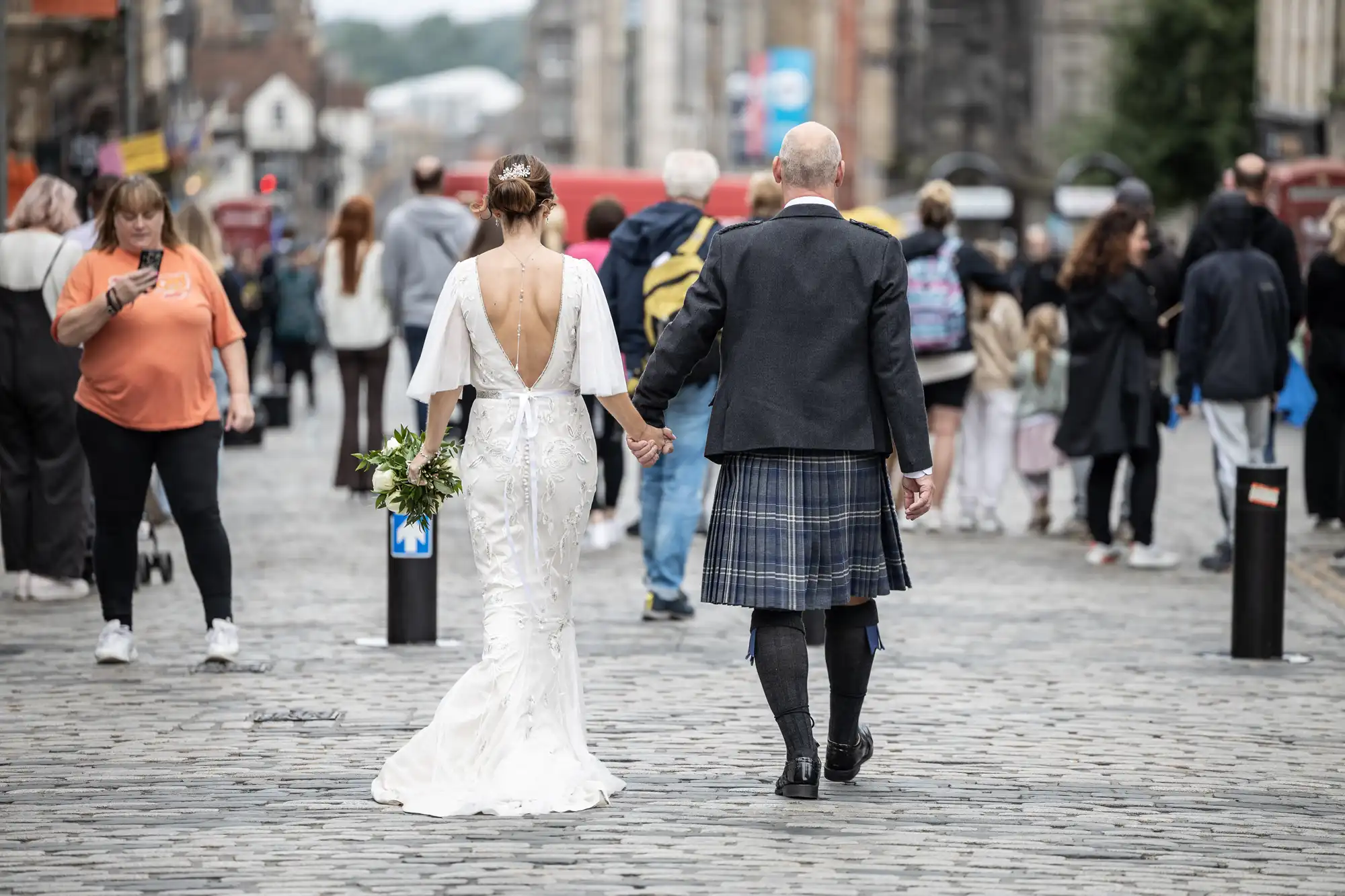 A bride in a white dress holding a bouquet and a man in traditional Scottish attire walk hand-in-hand on a cobblestone street, with people and buildings in the background.