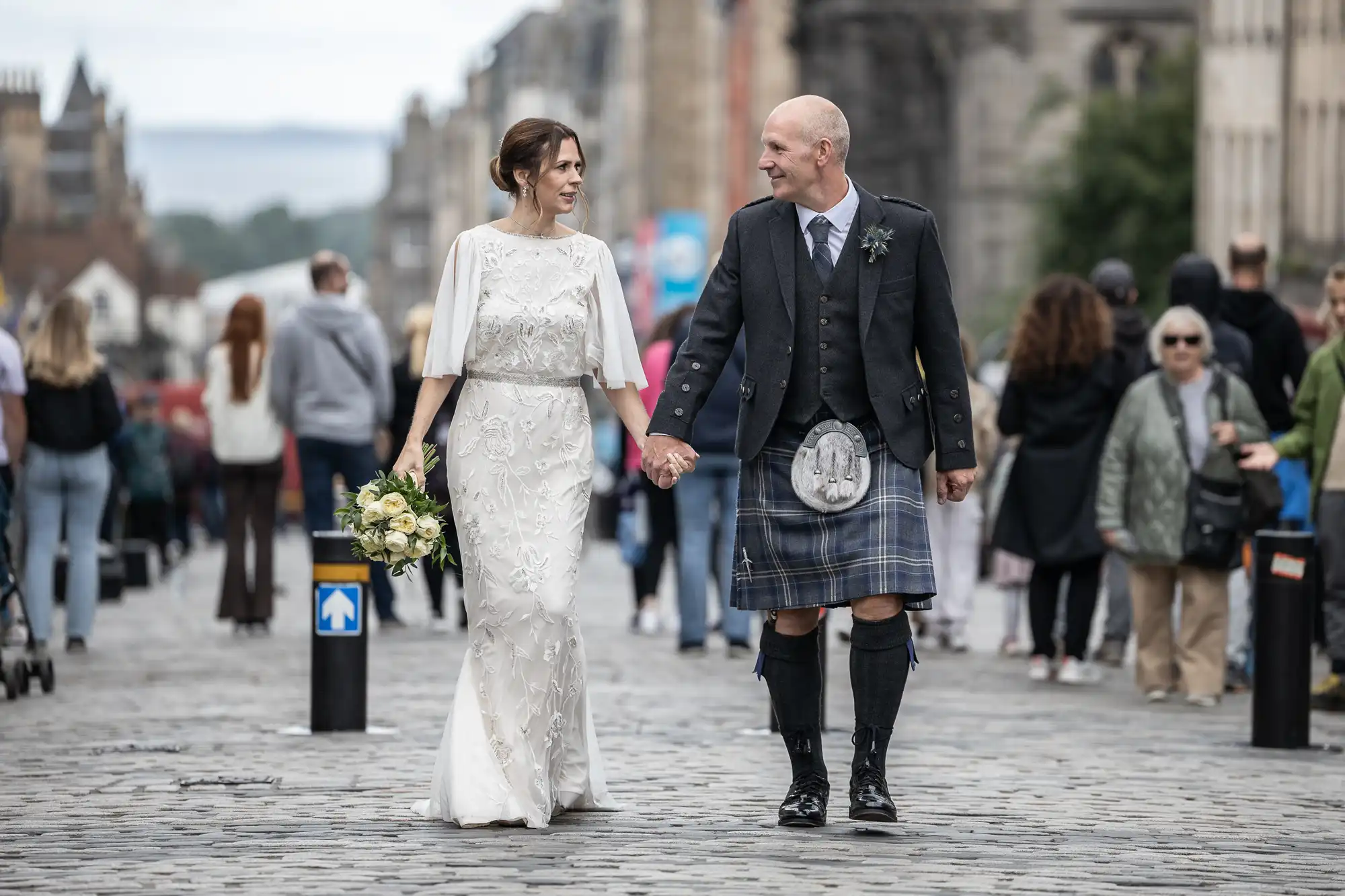 A couple, dressed in wedding attire, holds hands while walking down a cobblestone street surrounded by people. The man wears a kilt, and the woman carries a bouquet.