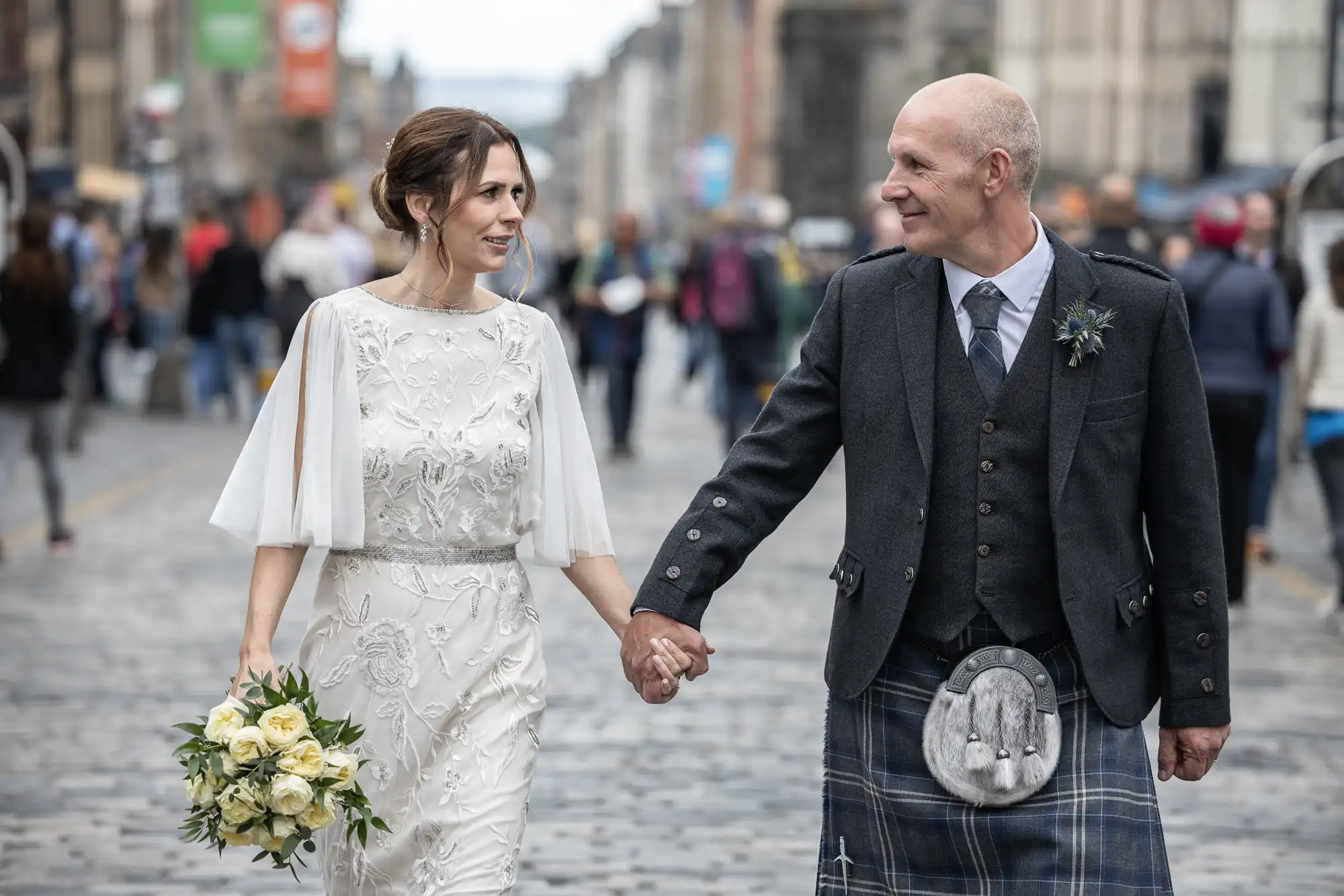 A couple, holding hands, walk down a cobblestone street dressed in wedding attire, with the man wearing a kilt and the woman holding a bouquet of flowers.