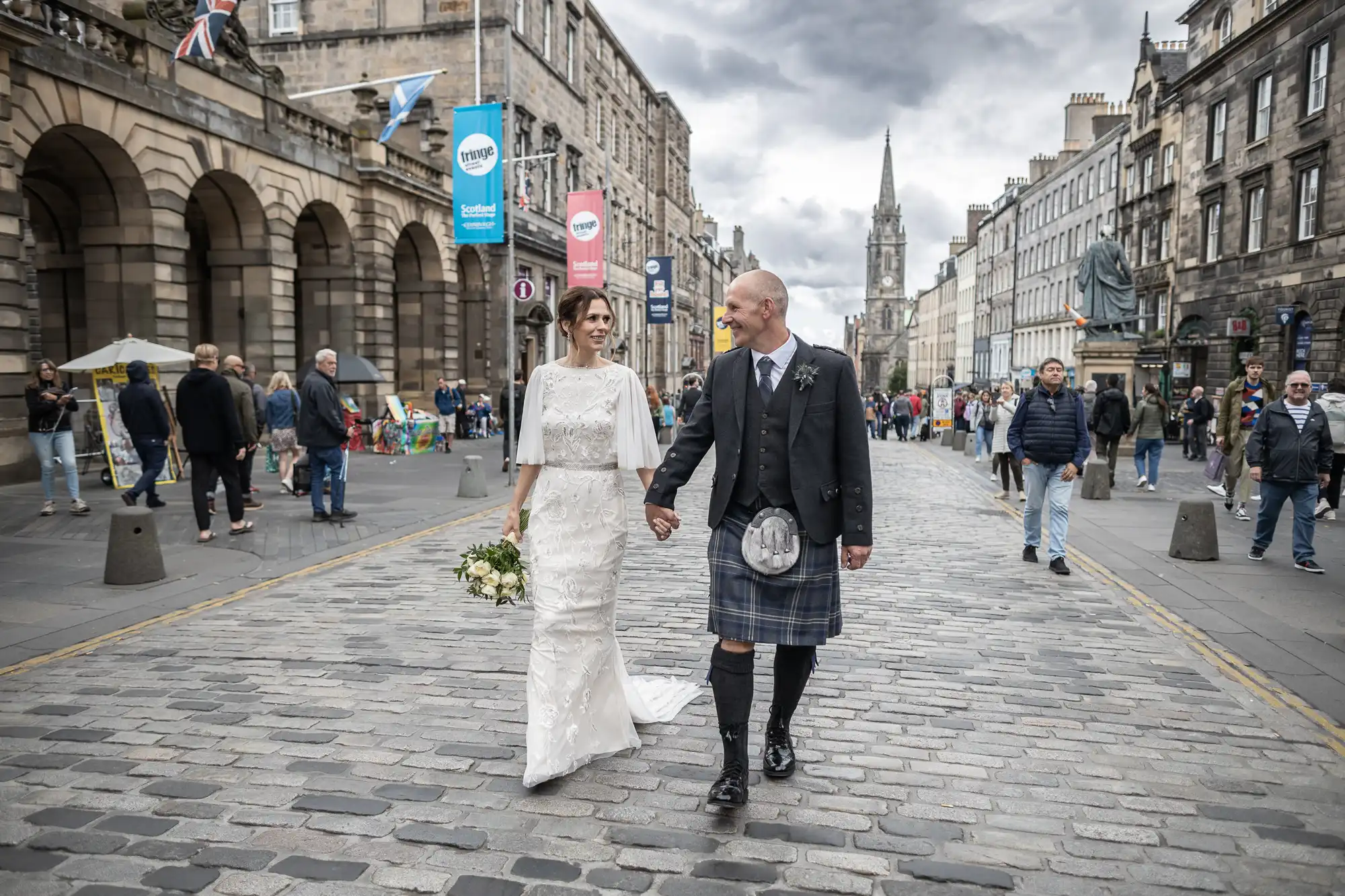 A bride and groom walk hand-in-hand along a cobbled street in a historic European city, surrounded by pedestrians and buildings under a cloudy sky.