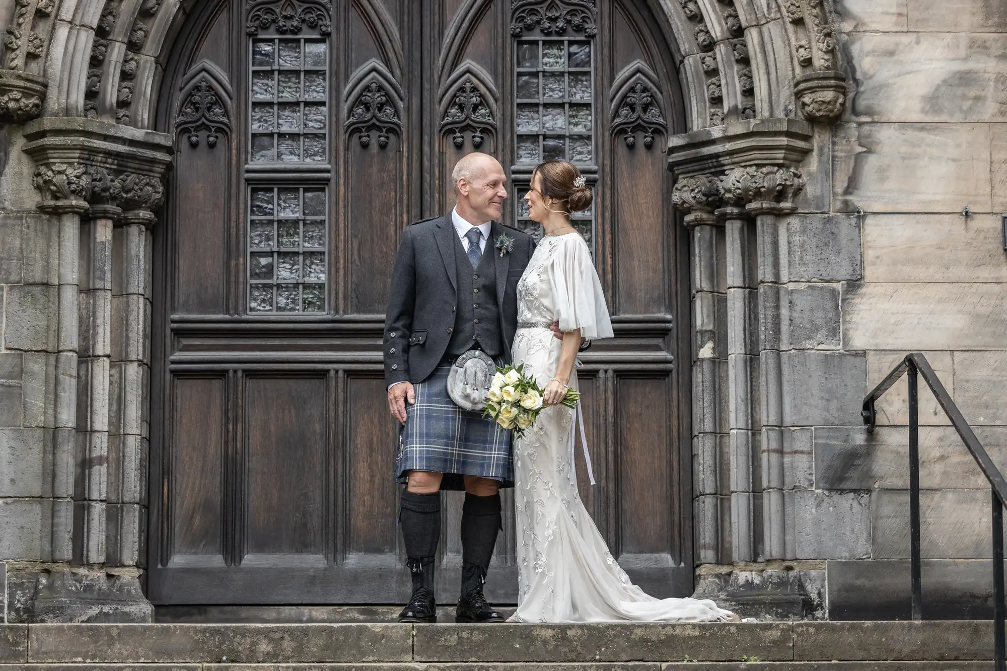 A couple stands in front of a decorative wooden door. The man is wearing a traditional kilt outfit, and the woman is wearing a white dress and holding a bouquet of flowers. They are looking at each other.