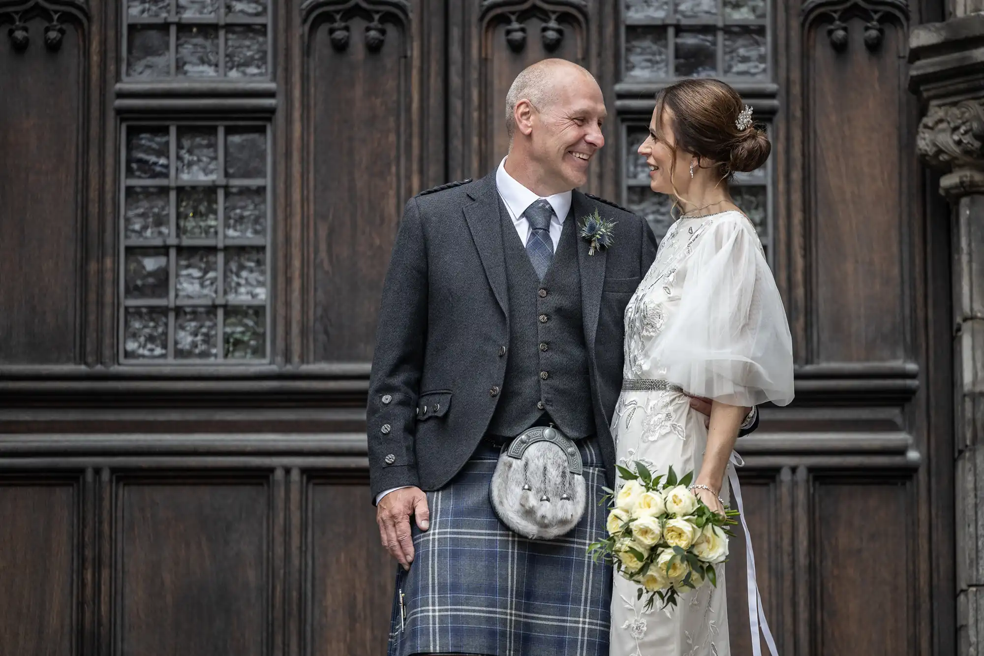 A couple dressed in formal attire, with the man in a kilt and jacket and the woman in a white dress holding a bouquet of flowers, smile at each other in front of a wooden door.