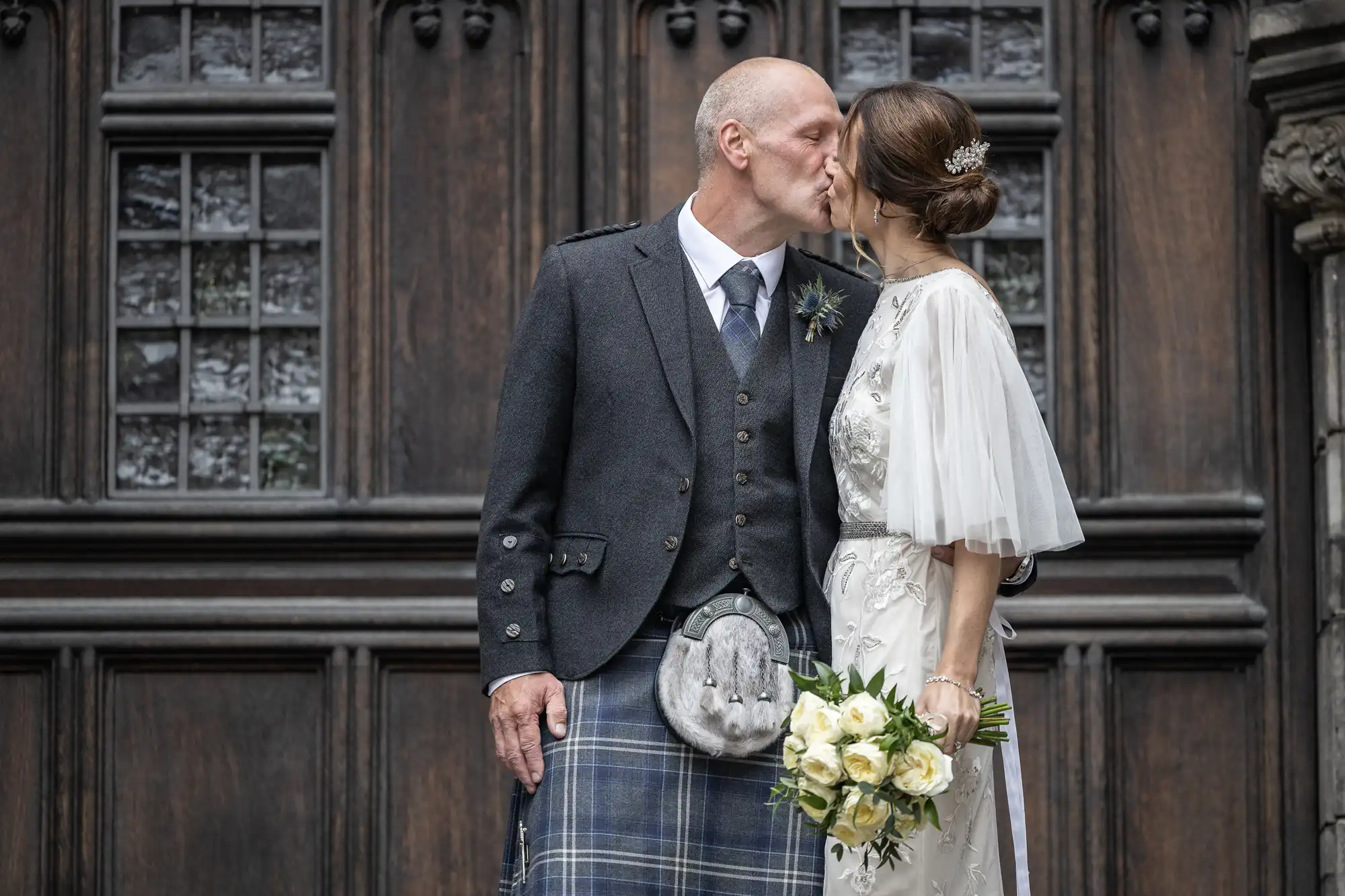 A couple in wedding attire kiss in front of a wooden door. The man wears a kilt and a grey jacket, and the woman wears a white dress and holds a bouquet of flowers.
