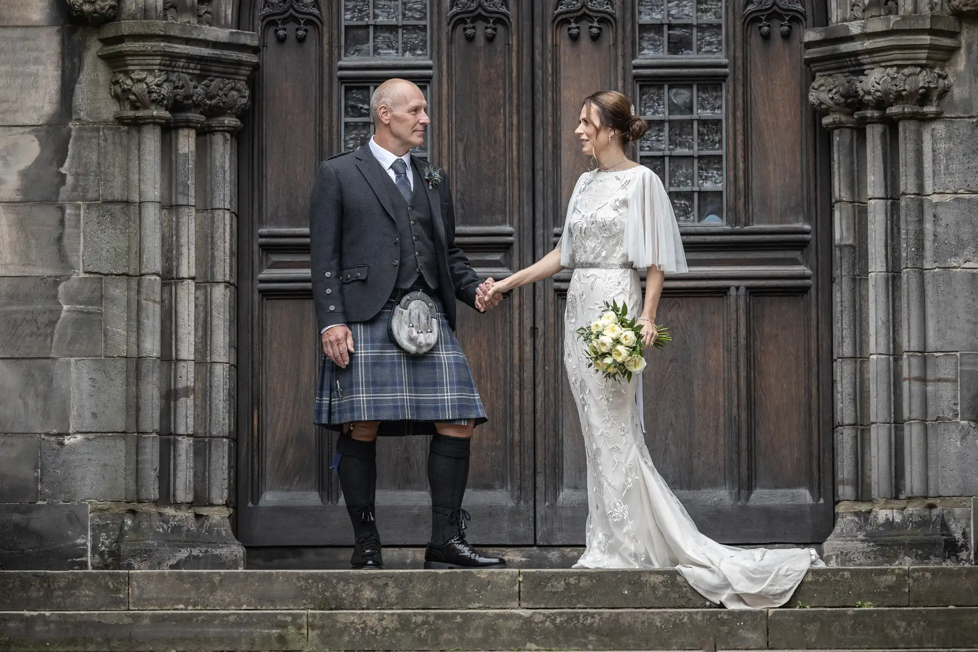 A man in traditional Scottish attire and a woman in a white dress hold hands on stone steps in front of a large wooden door with intricate carvings and tall windows.