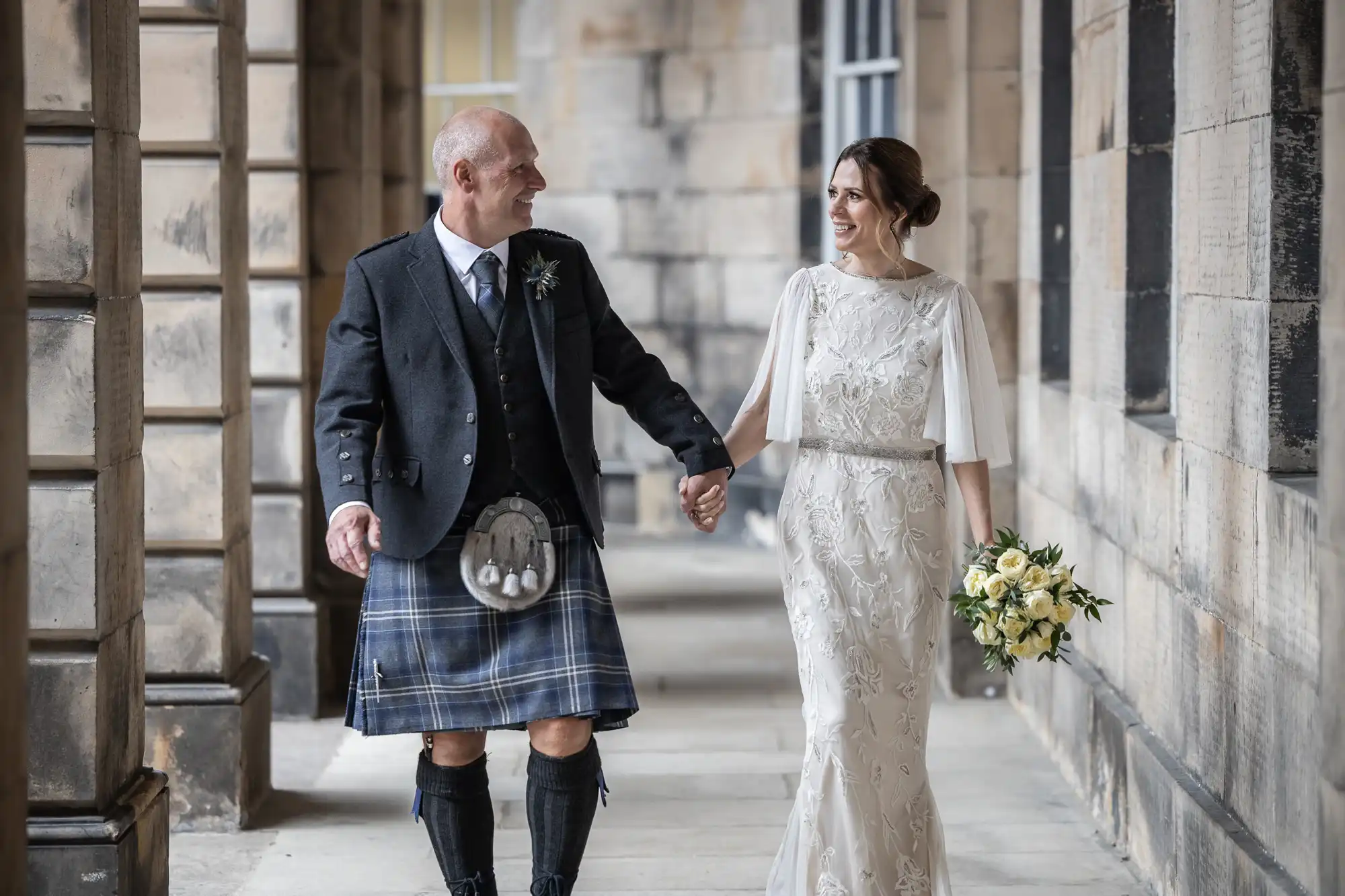 A couple, the man in a kilt and the woman in a white dress holding a bouquet, walk hand in hand in a stone corridor, both smiling at each other.