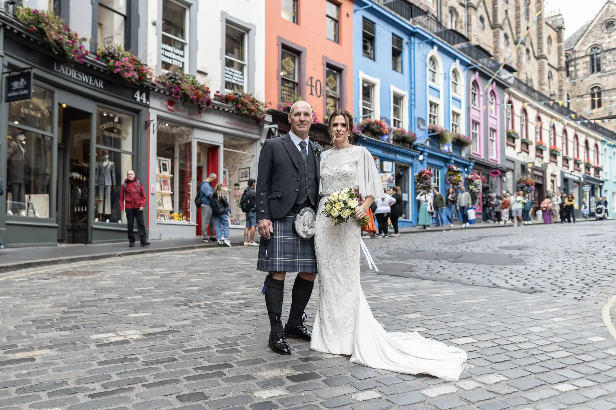 A couple dressed in wedding attire stands on a cobblestone street in front of a row of colorful buildings. The groom wears a kilt, and the bride holds a bouquet. People can be seen in the background.