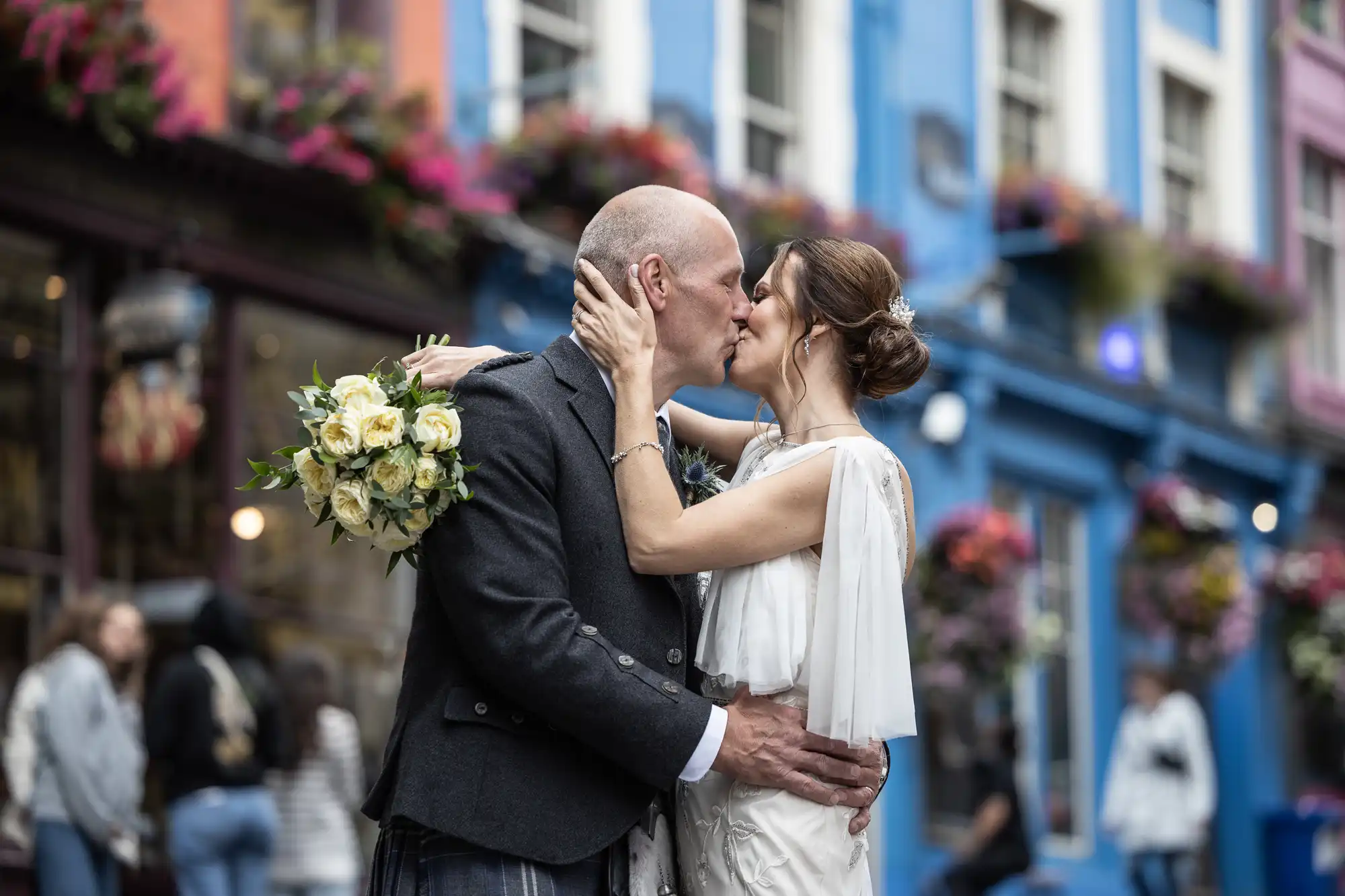 A couple in wedding attire shares a kiss on a colorful street. The groom holds a bouquet of flowers.