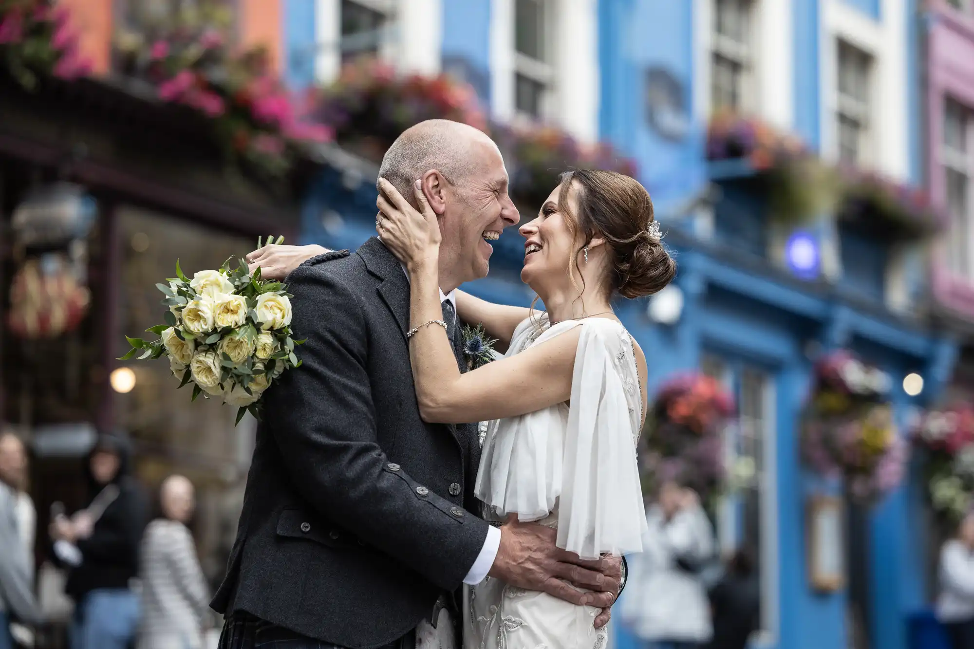A couple in wedding attire embraces and smiles at each other while standing on a colorful street adorned with flowers.