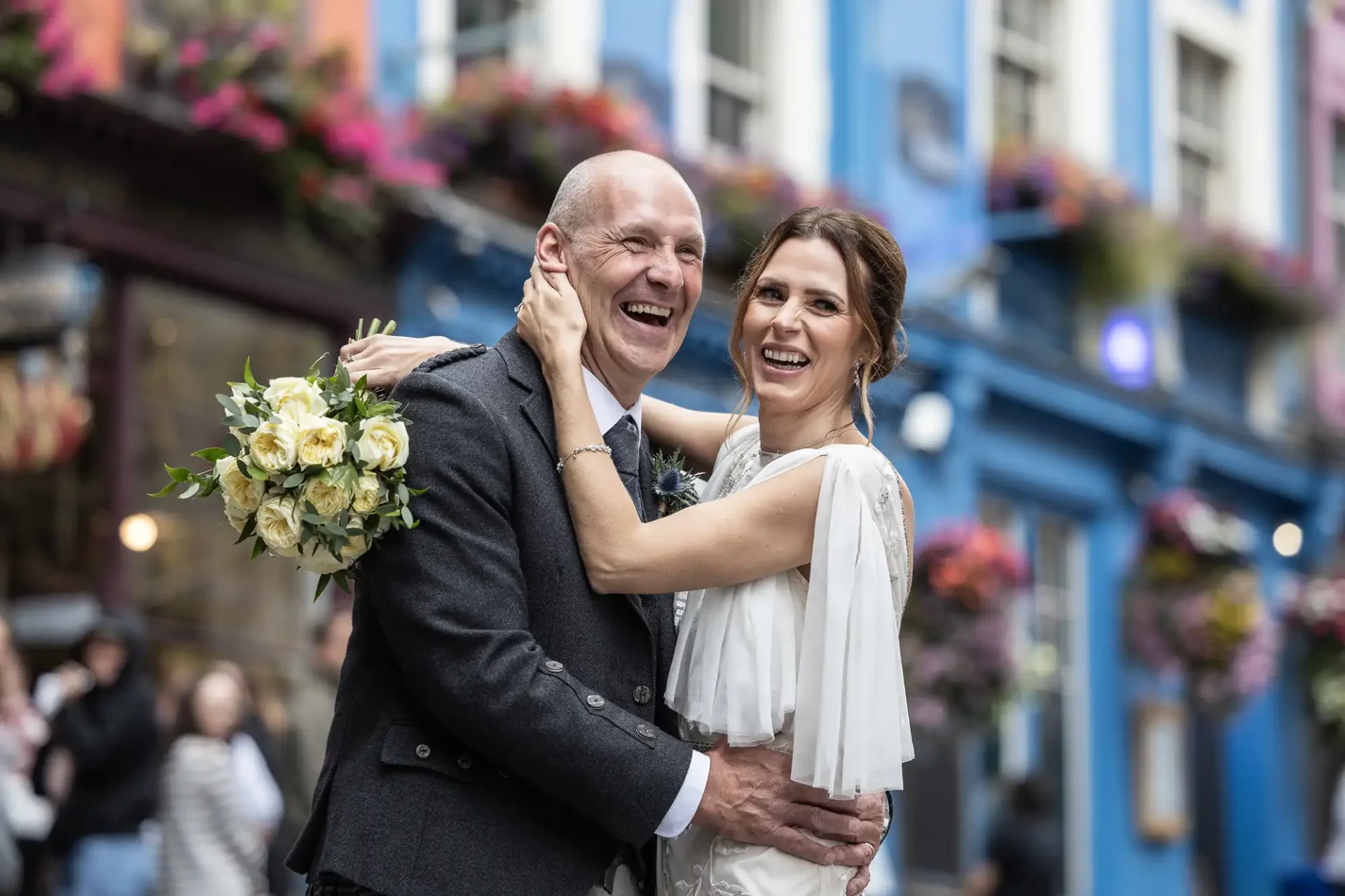 A smiling couple in formal attire, with the woman holding a bouquet of flowers, poses in front of a colorful, flower-adorned building.