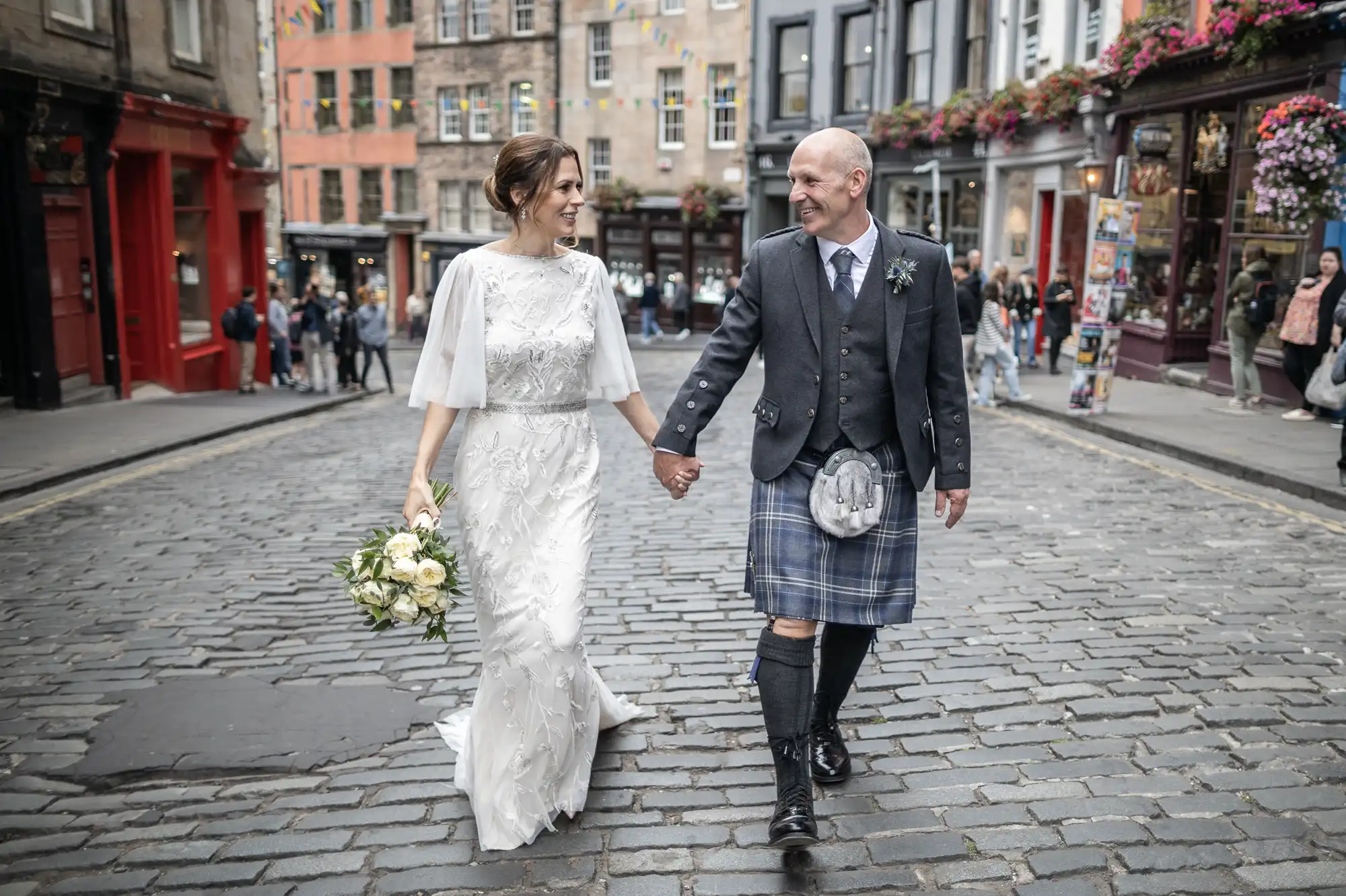 A couple, with the man in a kilt and the woman in a white wedding dress, walk hand-in-hand on a cobblestone street in a city setting. The man holds a bouquet of flowers.