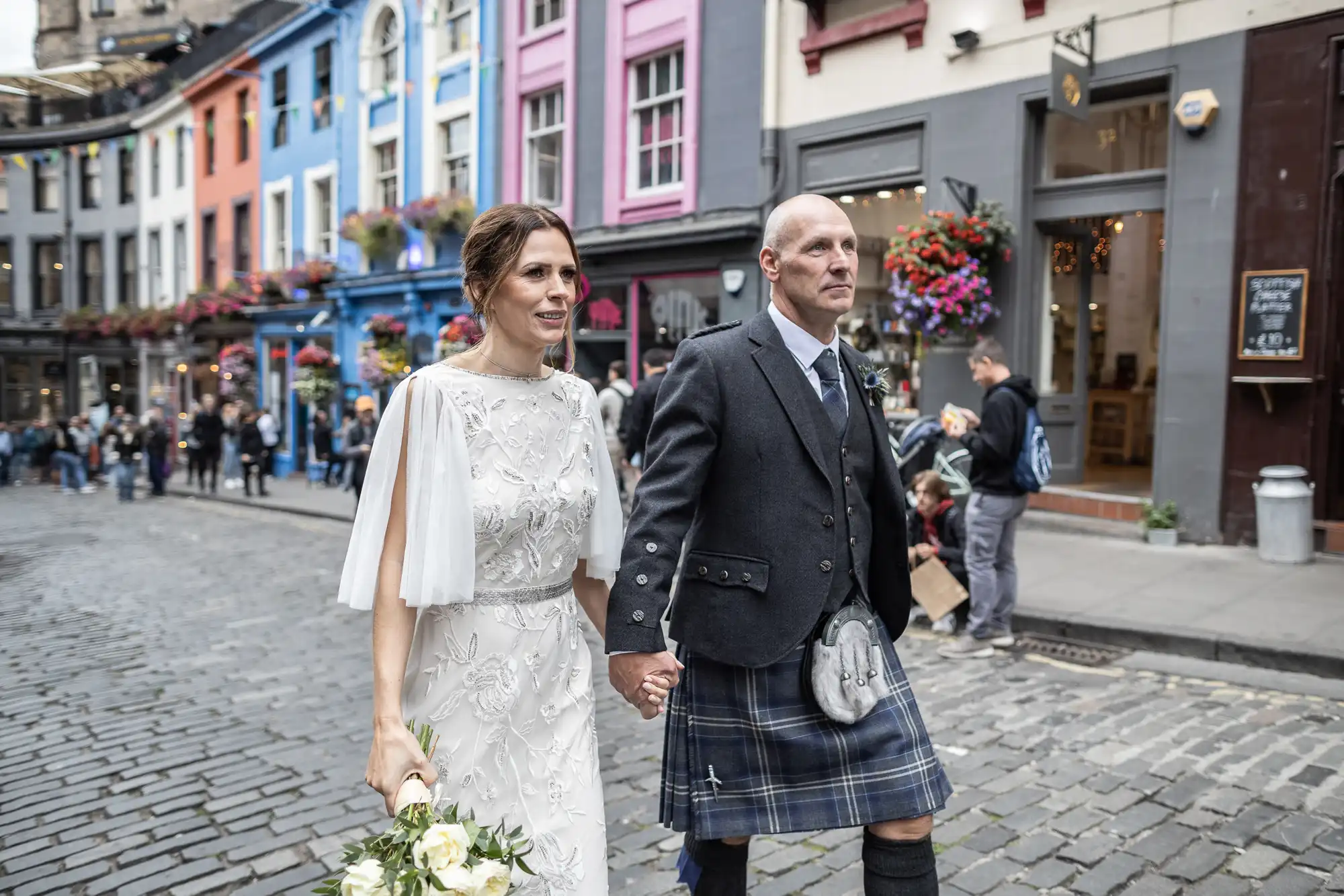 A couple dressed in wedding attire walks hand in hand on a cobblestone street lined with colorful buildings and shops. The woman wears a white dress, and the man wears a kilt and suit jacket.