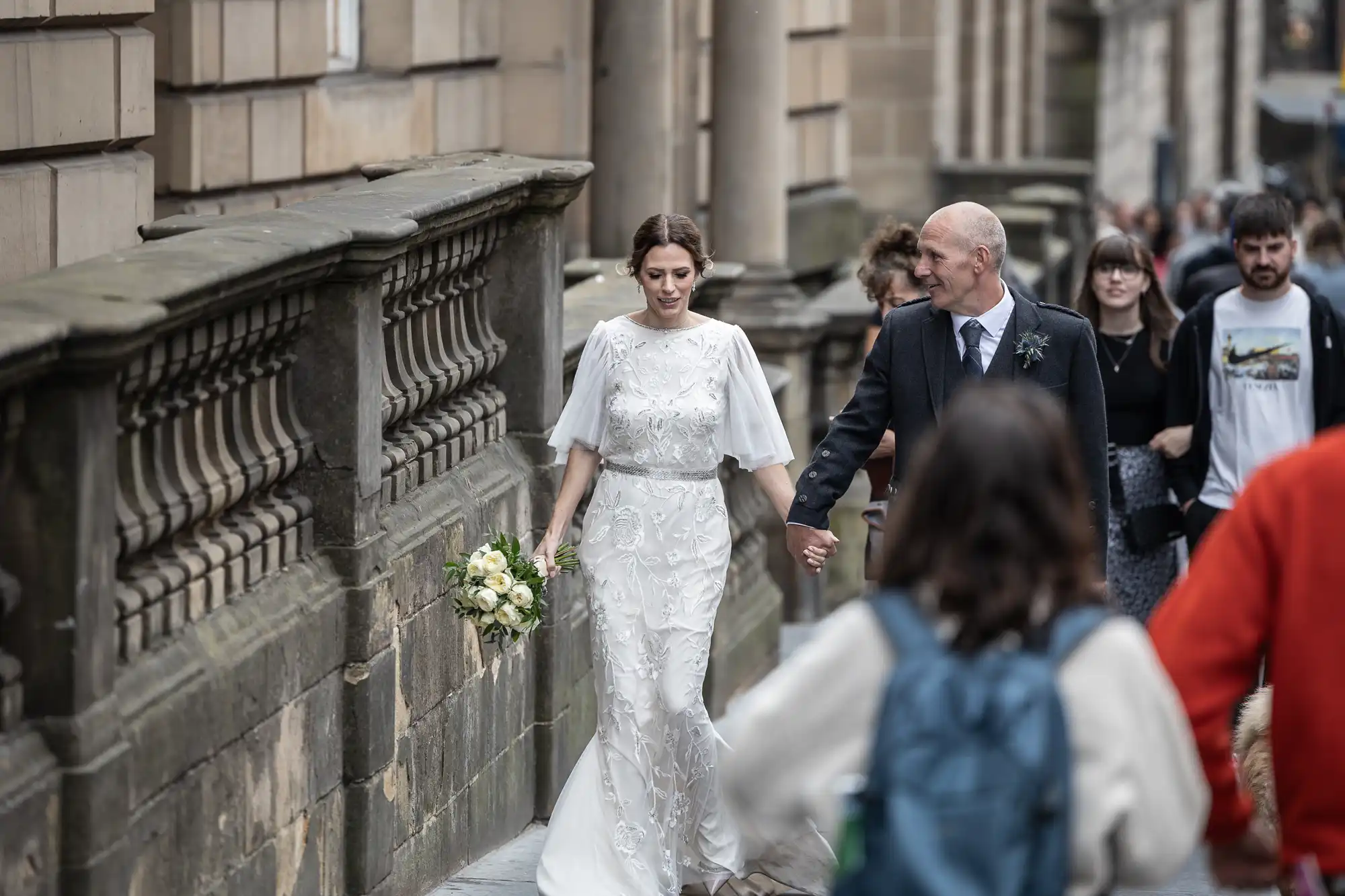 A bride in a white gown holding a bouquet walks beside a man in a suit, with people walking nearby on a stone-paved street near ornate stone buildings.