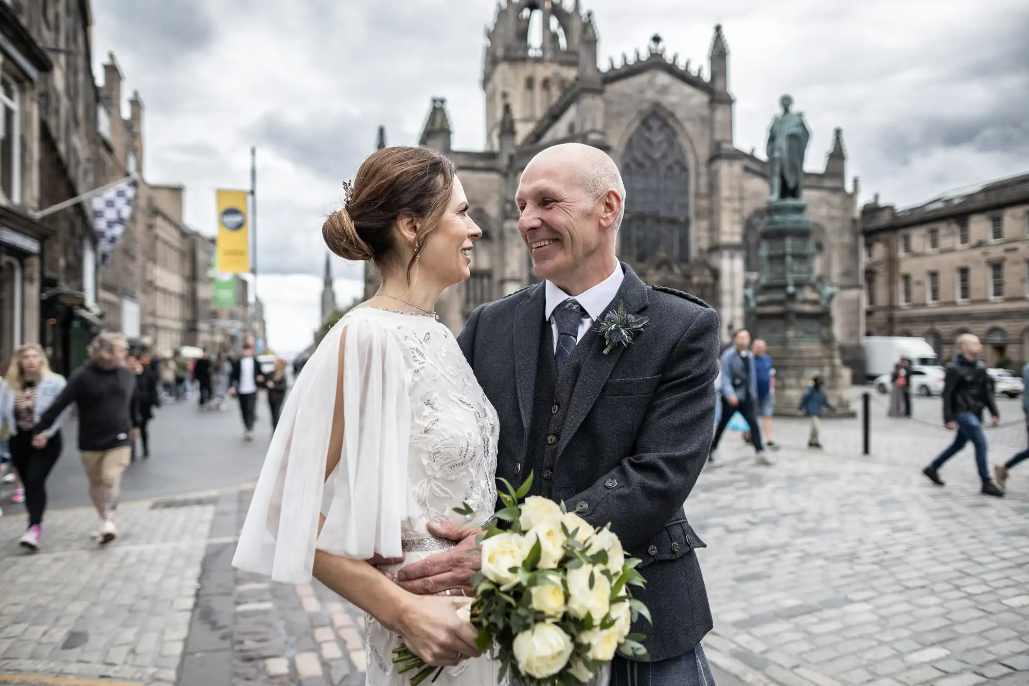 A smiling couple in wedding attire stands closely together holding a bouquet on a cobblestone street, with a historic church and people in the background.
