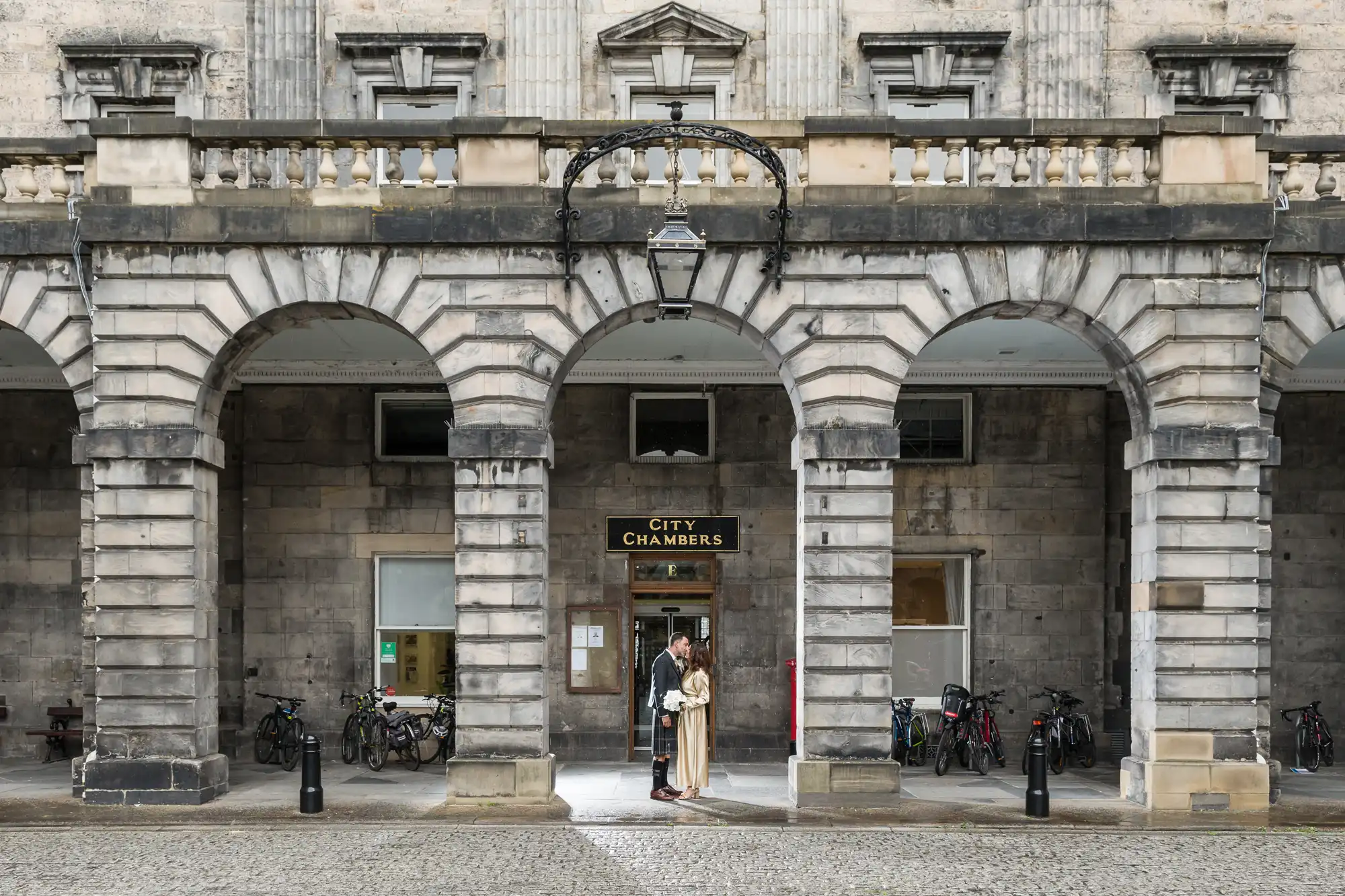 Newlyweds outside a building labelled "City Chambers," with several bicycles parked nearby and an arched stone façade in the background.