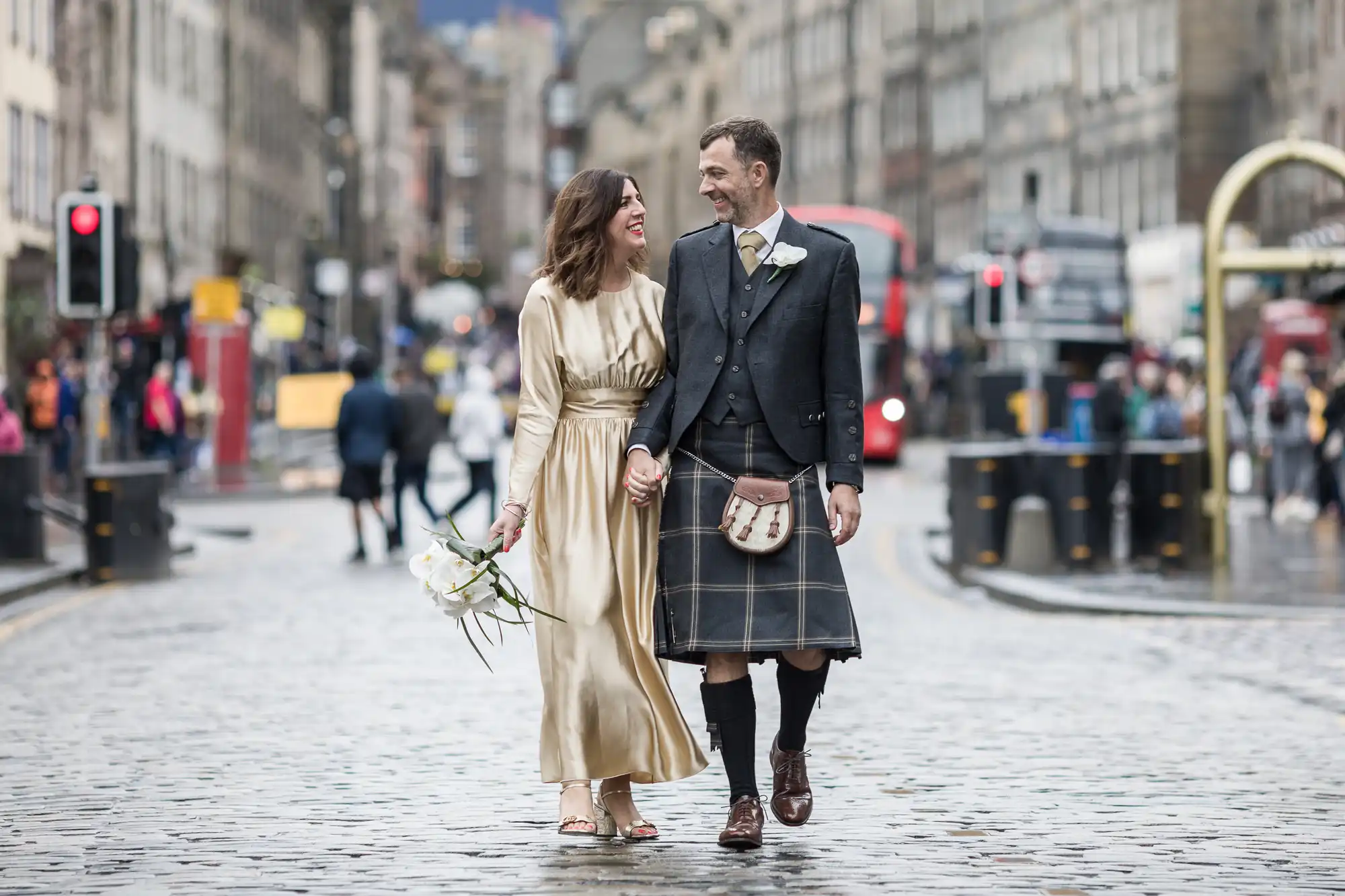 A newlywed couple dressed in formal attire, with the man wearing a kilt, walk hand in hand on a cobblestone street in a city. The woman holds a bouquet of white flowers.