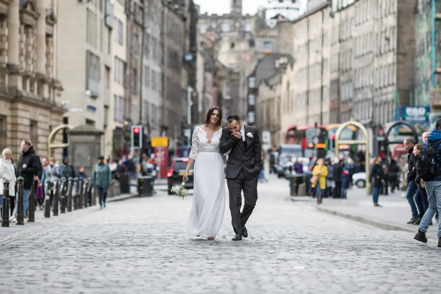 A bride and groom walk arm-in-arm down a cobblestone street in a city surrounded by buildings and people.