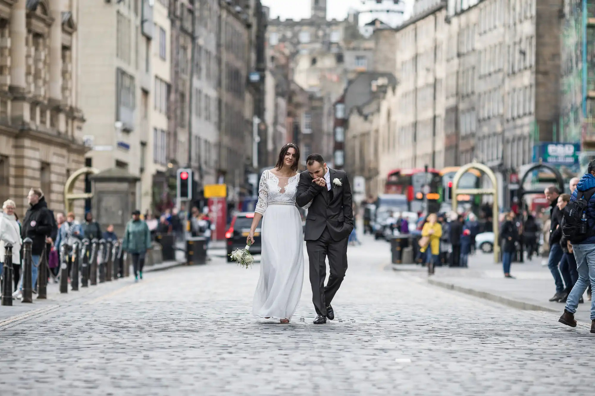 A bride and groom walk arm-in-arm down a cobblestone street in a city surrounded by buildings and people.