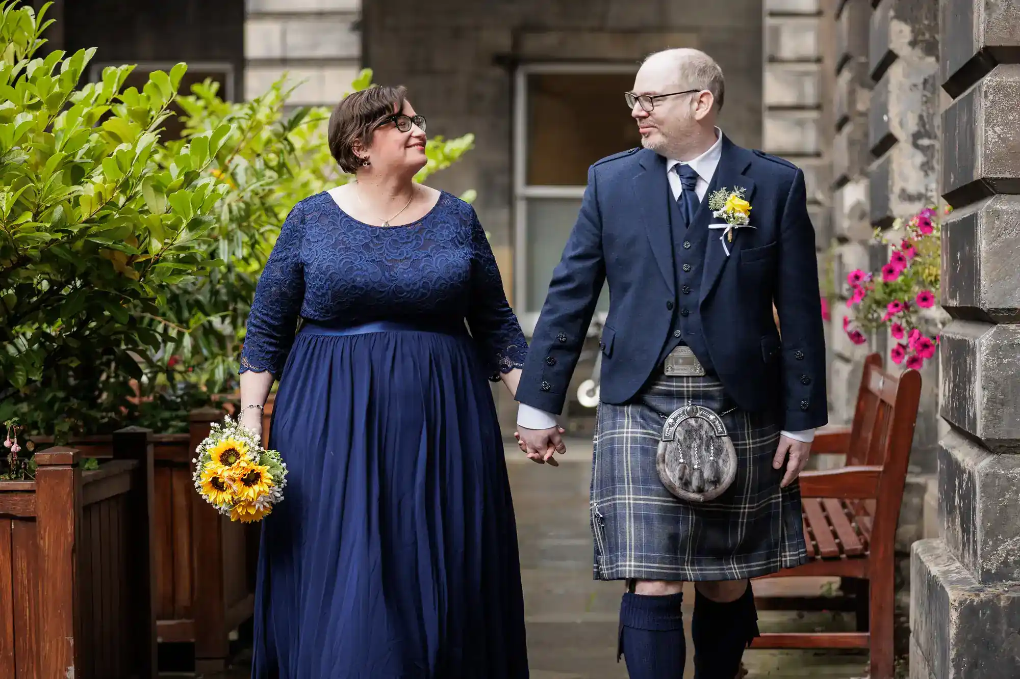 A couple holds hands while walking outdoors; the woman wears a long blue dress and holds sunflowers, and the man wears a blue suit jacket and tartan kilt.